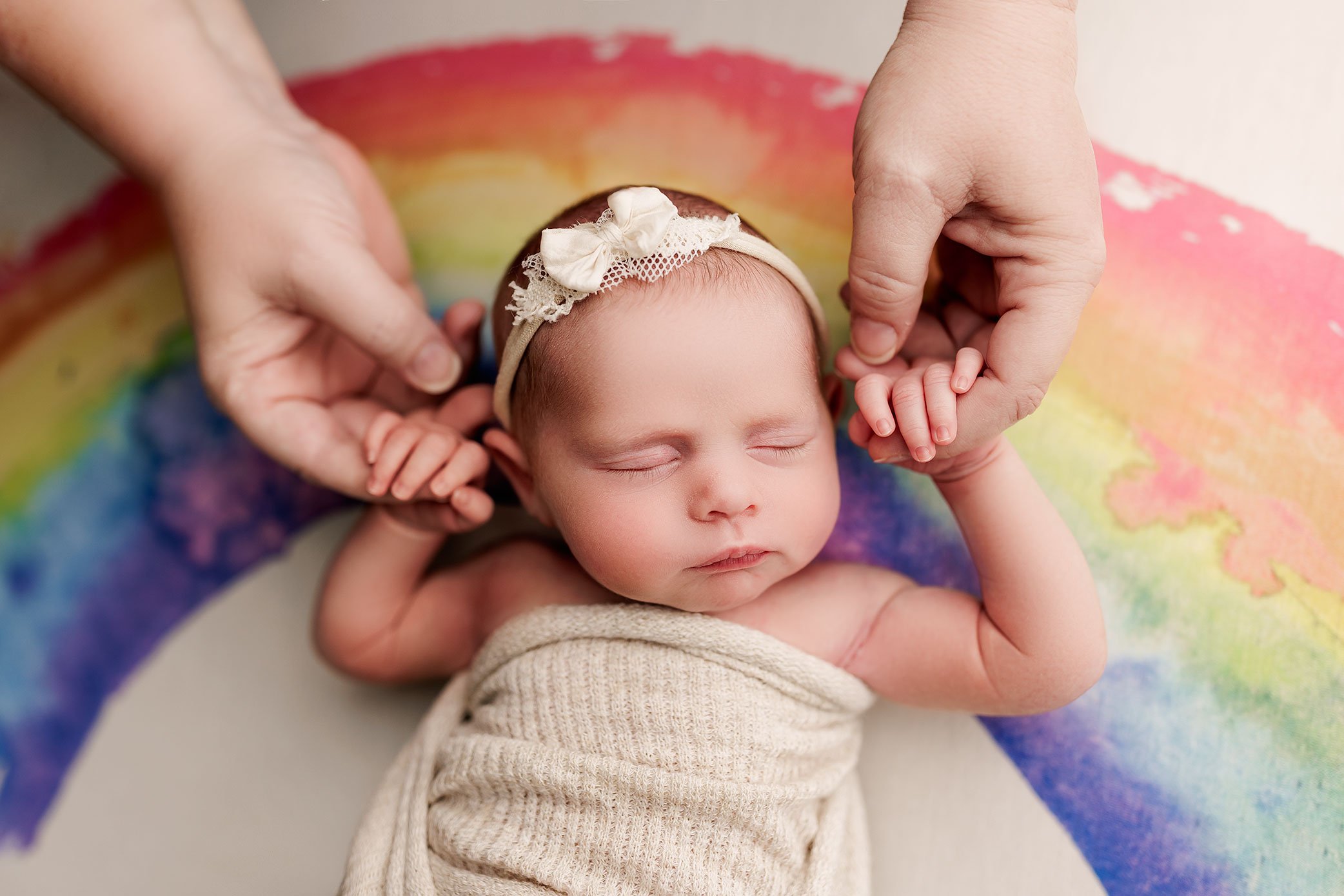 Sleeping newborn baby girl on rainbow backdrop gently holding her mom's hands.