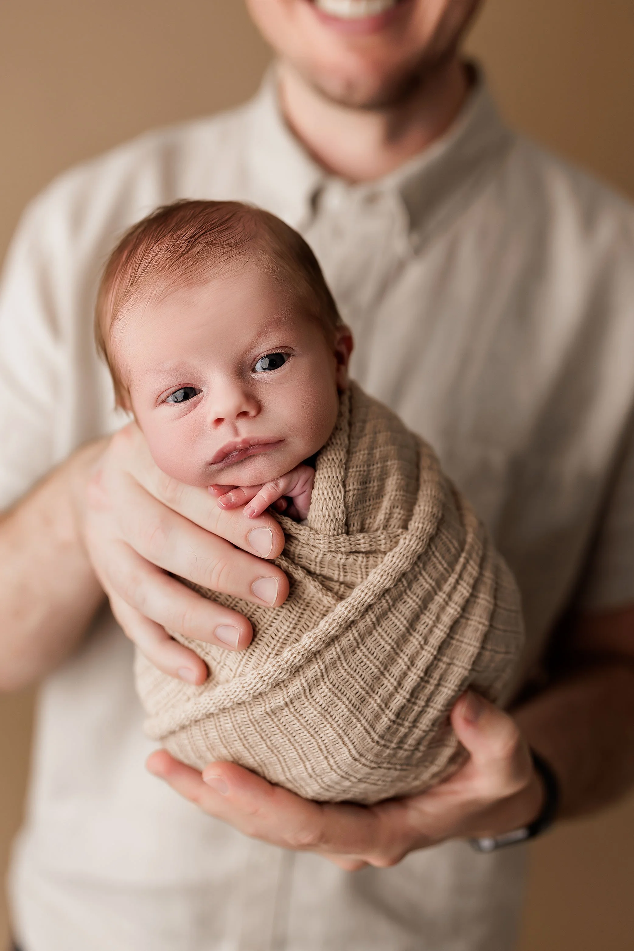 Awake newborn baby boy wrapped in a neutral knit swaddle, held securely in his father’s arms.