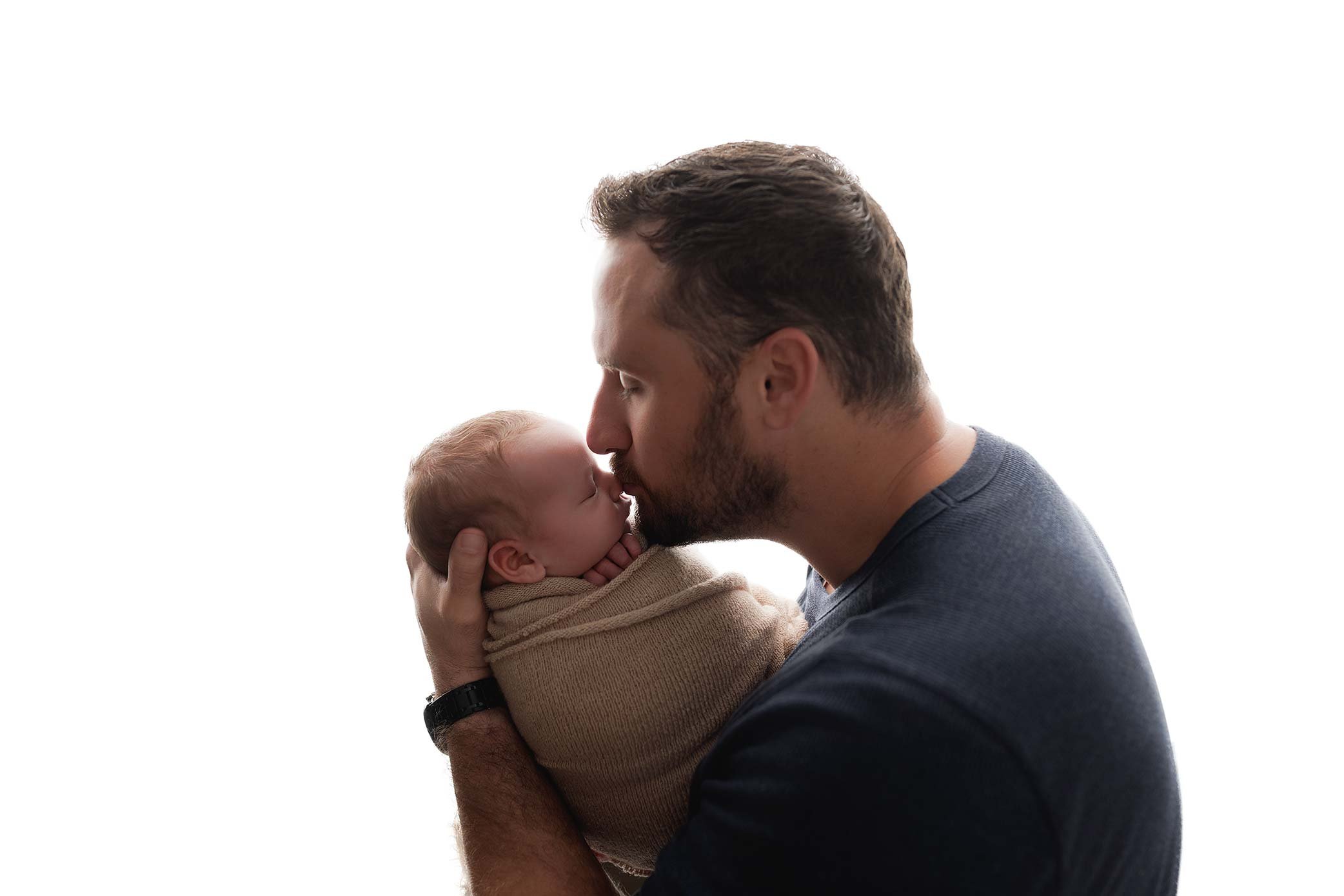 Father gently kissing his swaddled newborn baby on the nose in a soft, backlit studio portrait.