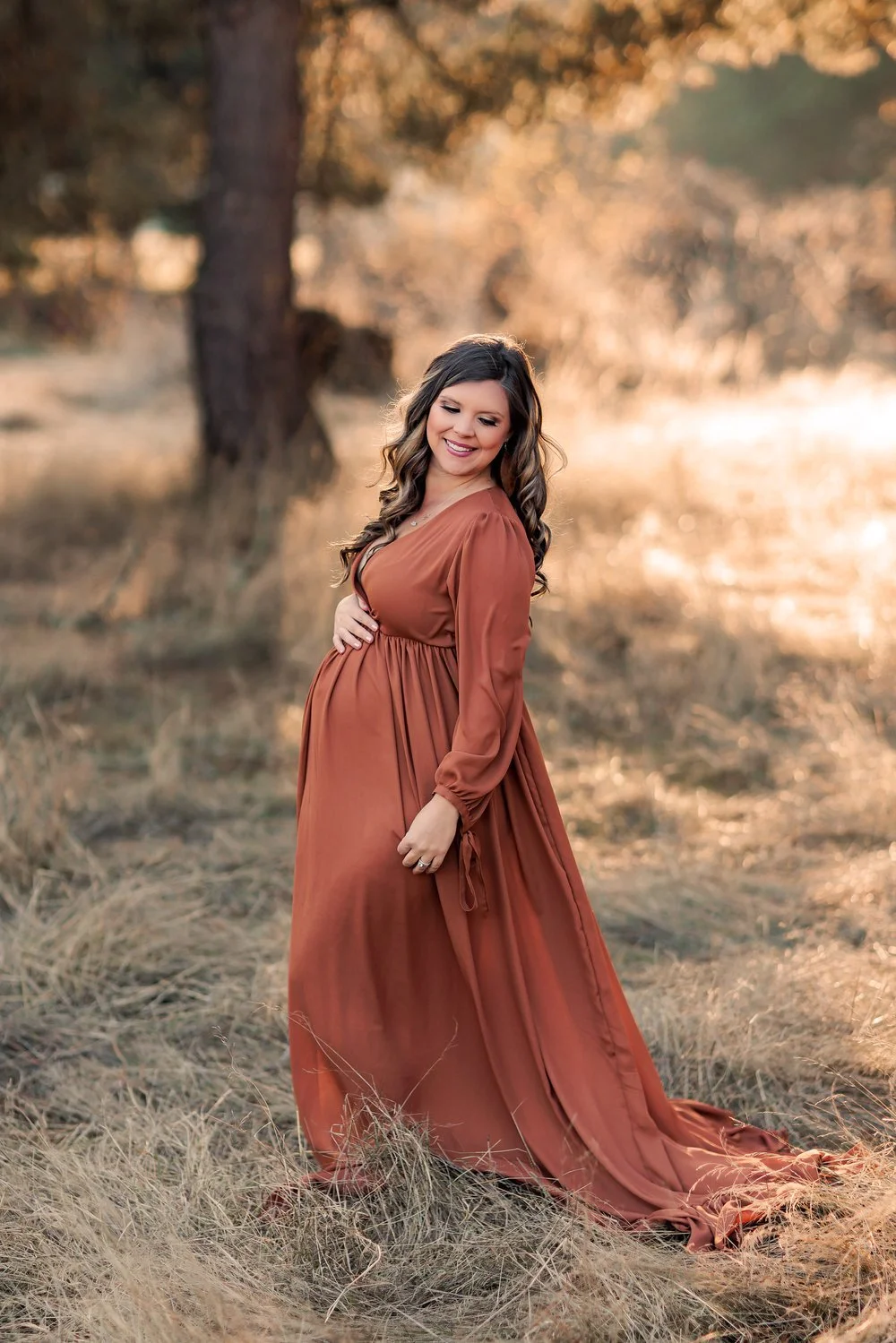 Pregnant mother in flowing rust dress standing in golden field during maternity session.