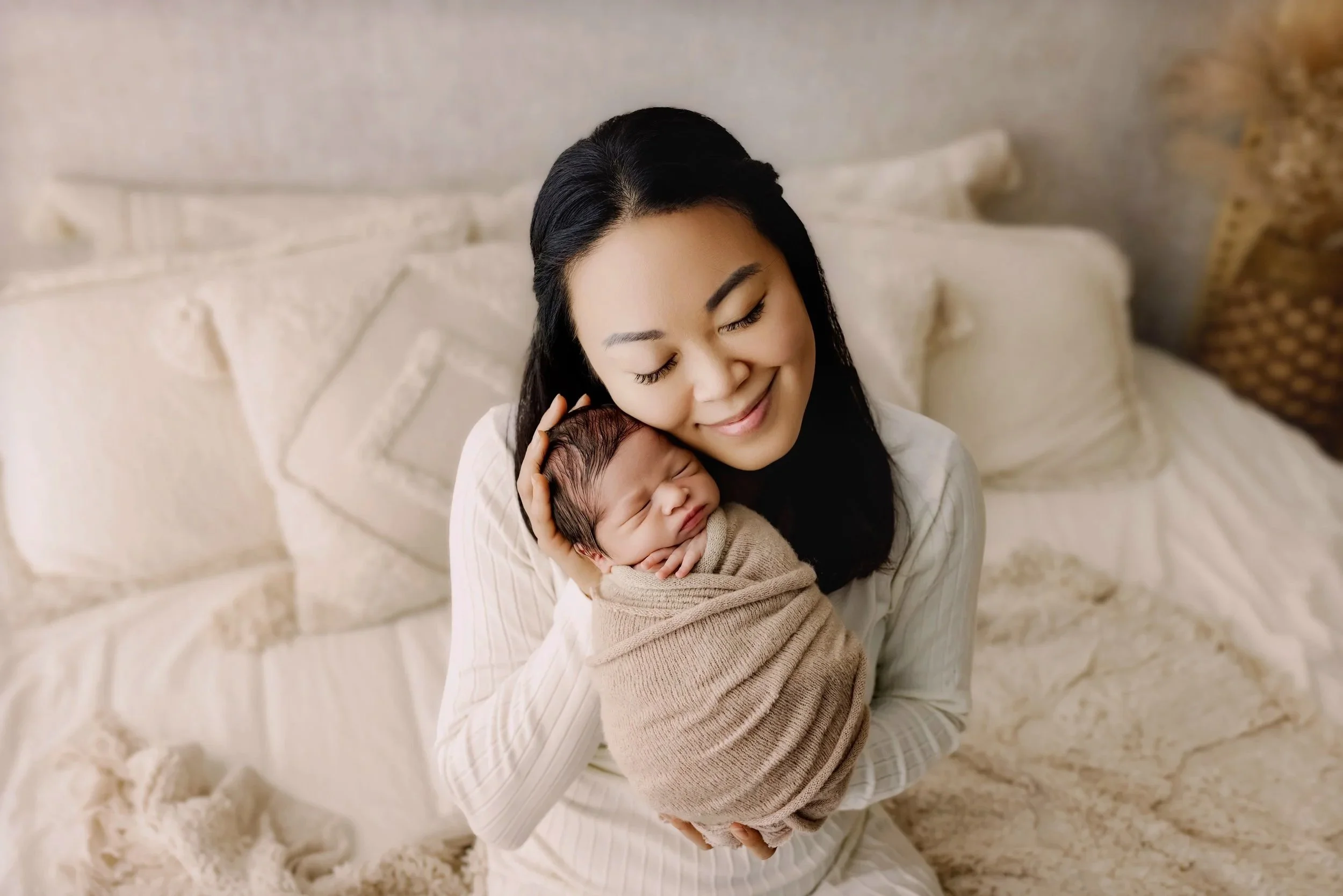 Mother gently cradling her sleeping newborn baby wrapped in neutral fabric in cozy bedroom setting.