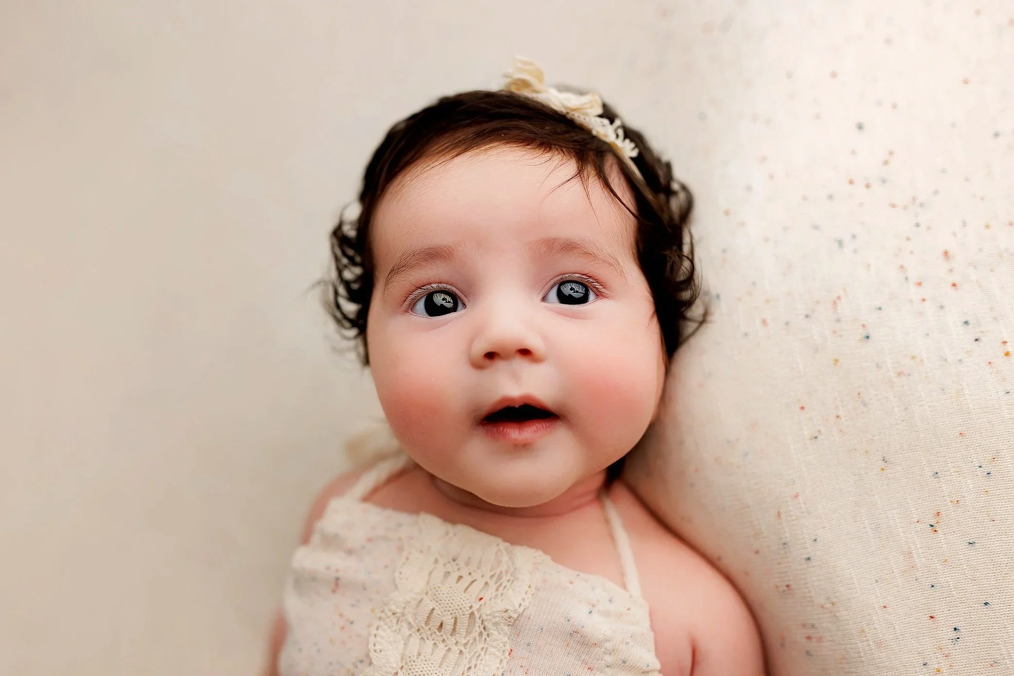 Baby girl with dark curly hair wearing cream lace outfit looking up during milestone session.