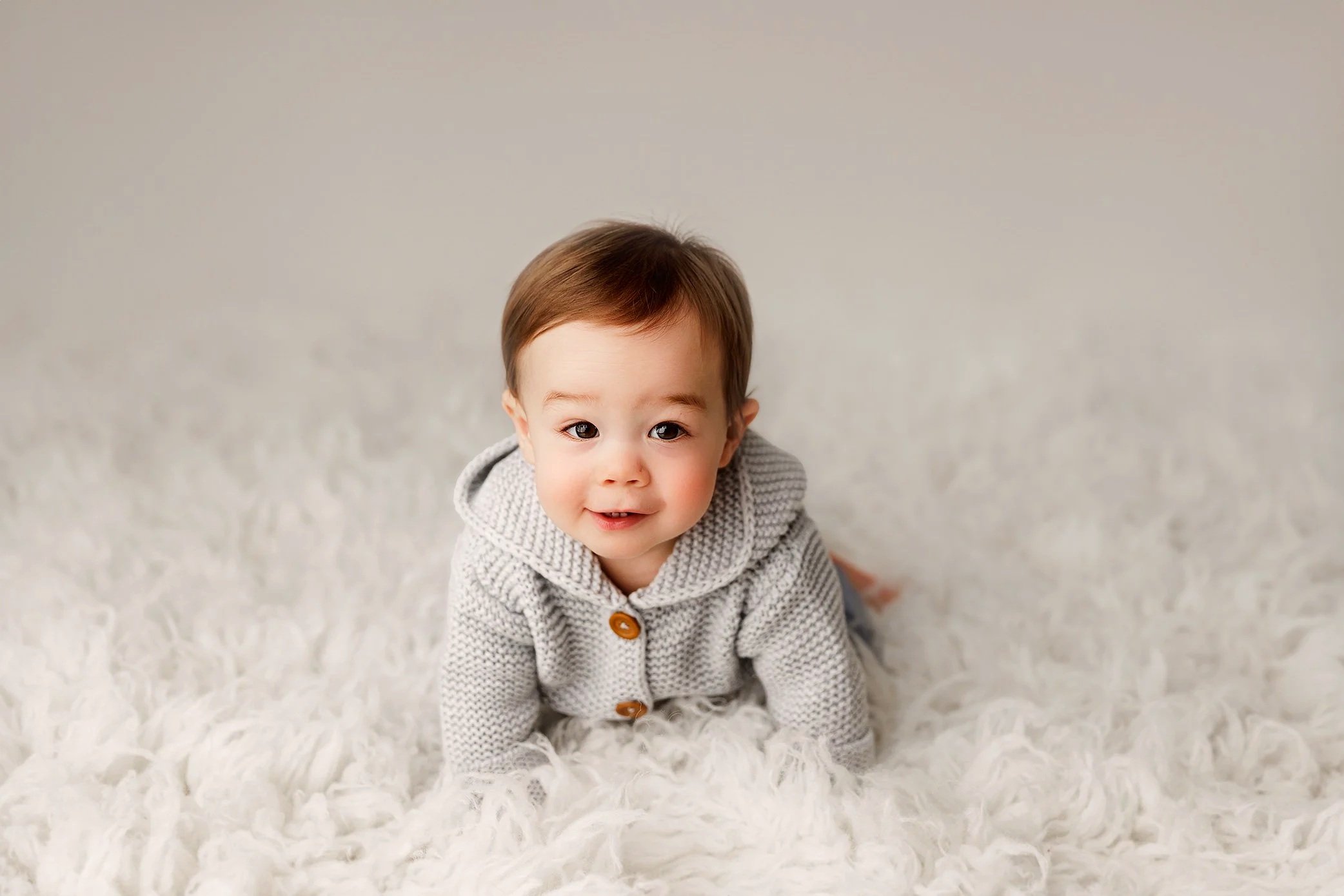 Six month old baby boy wearing grey knit sweater lying on soft white blanket.