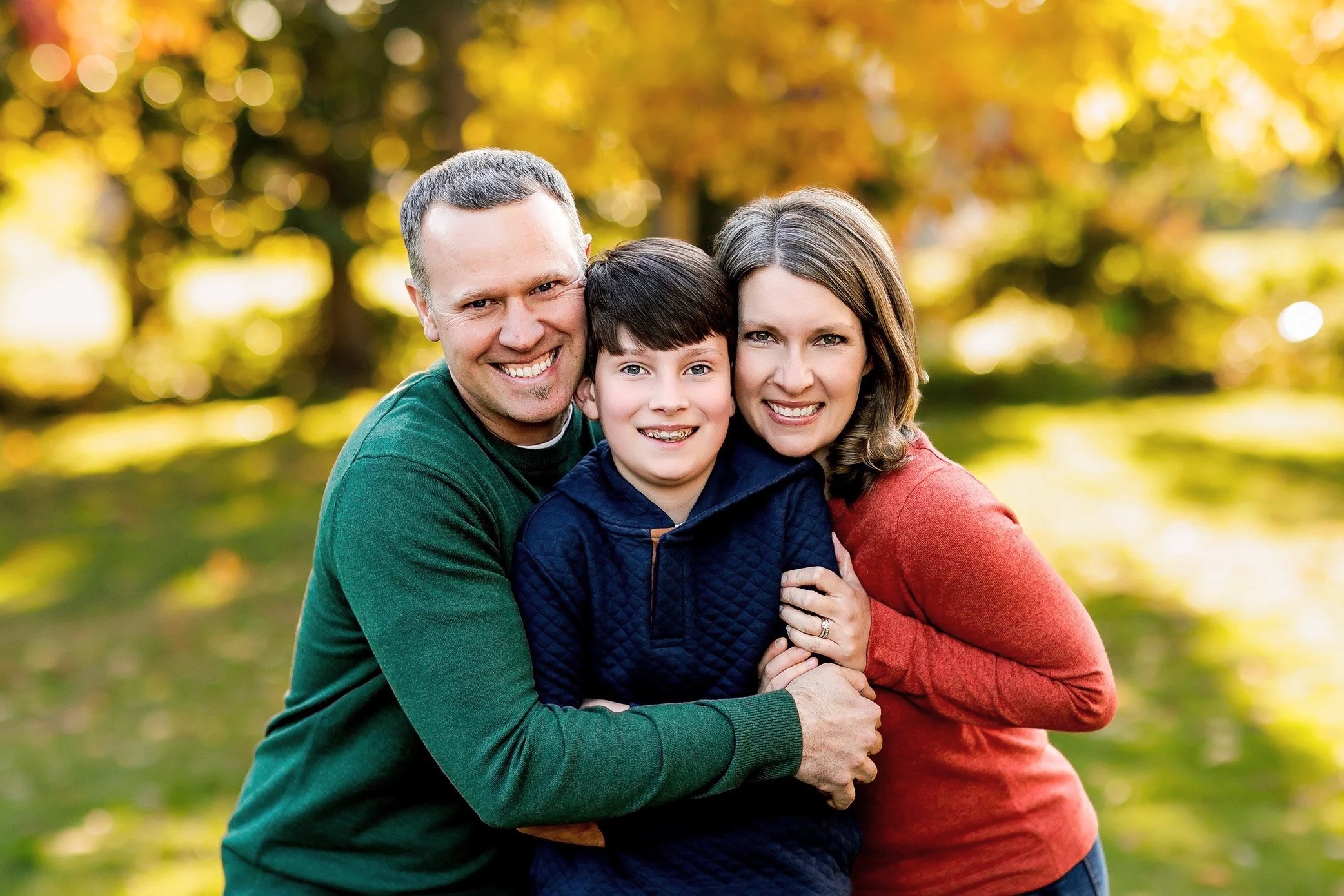 Family of three close together with child in the middle as parents hug tightly in park.
