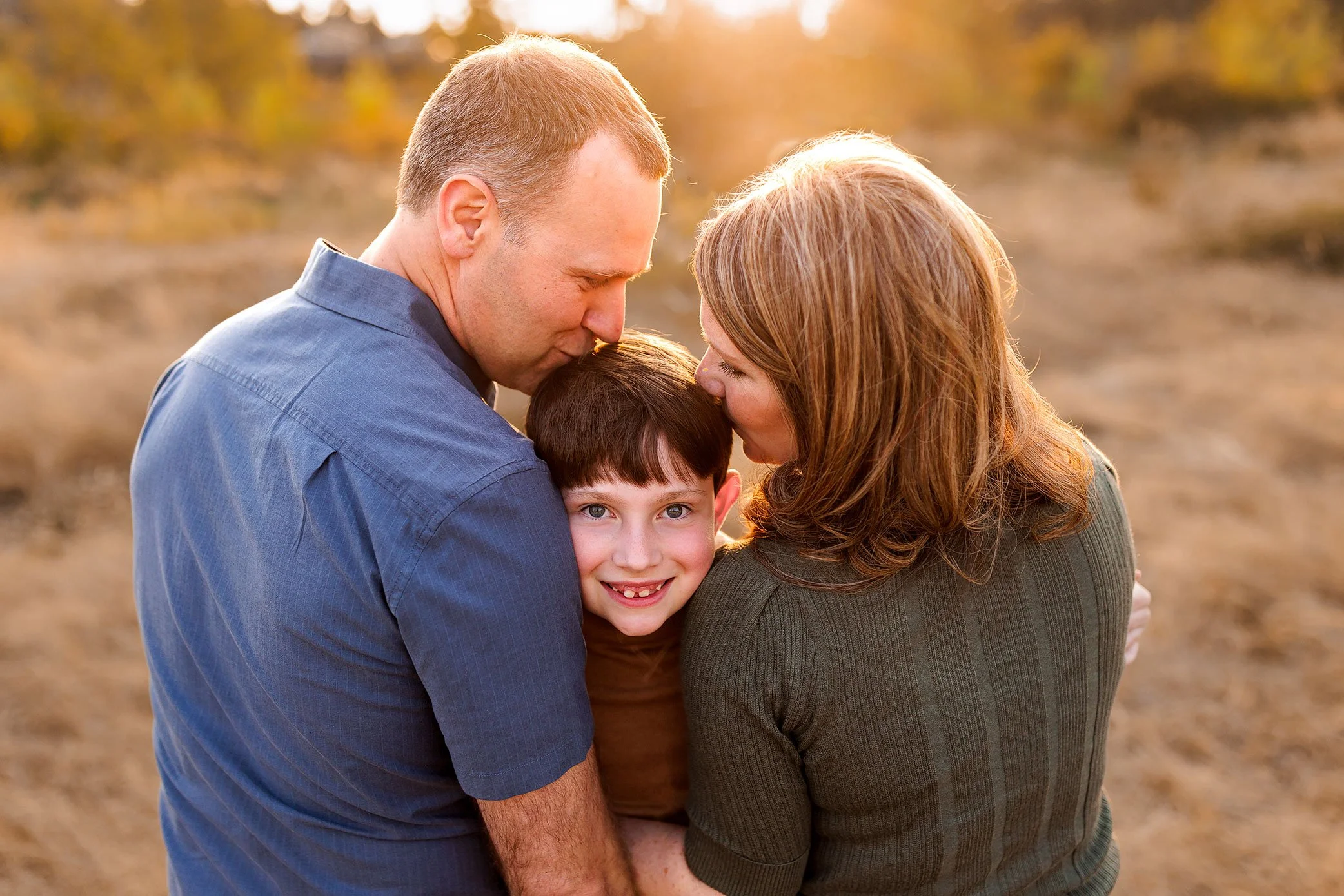Mother and father kiss their smiling son on the forehead in a warm sunset field portrait.