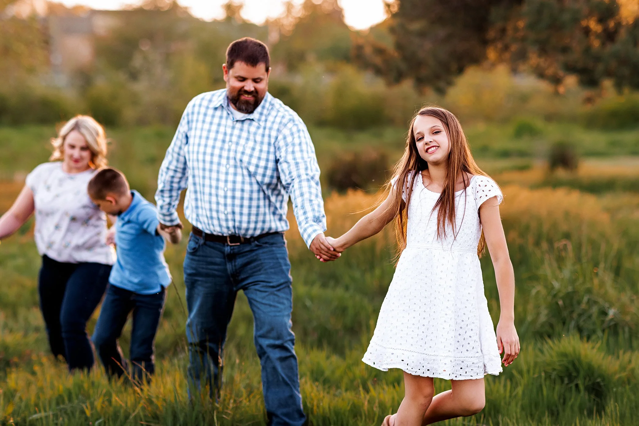 Family of four walking through grassy field with daughter leading parents toward camera.