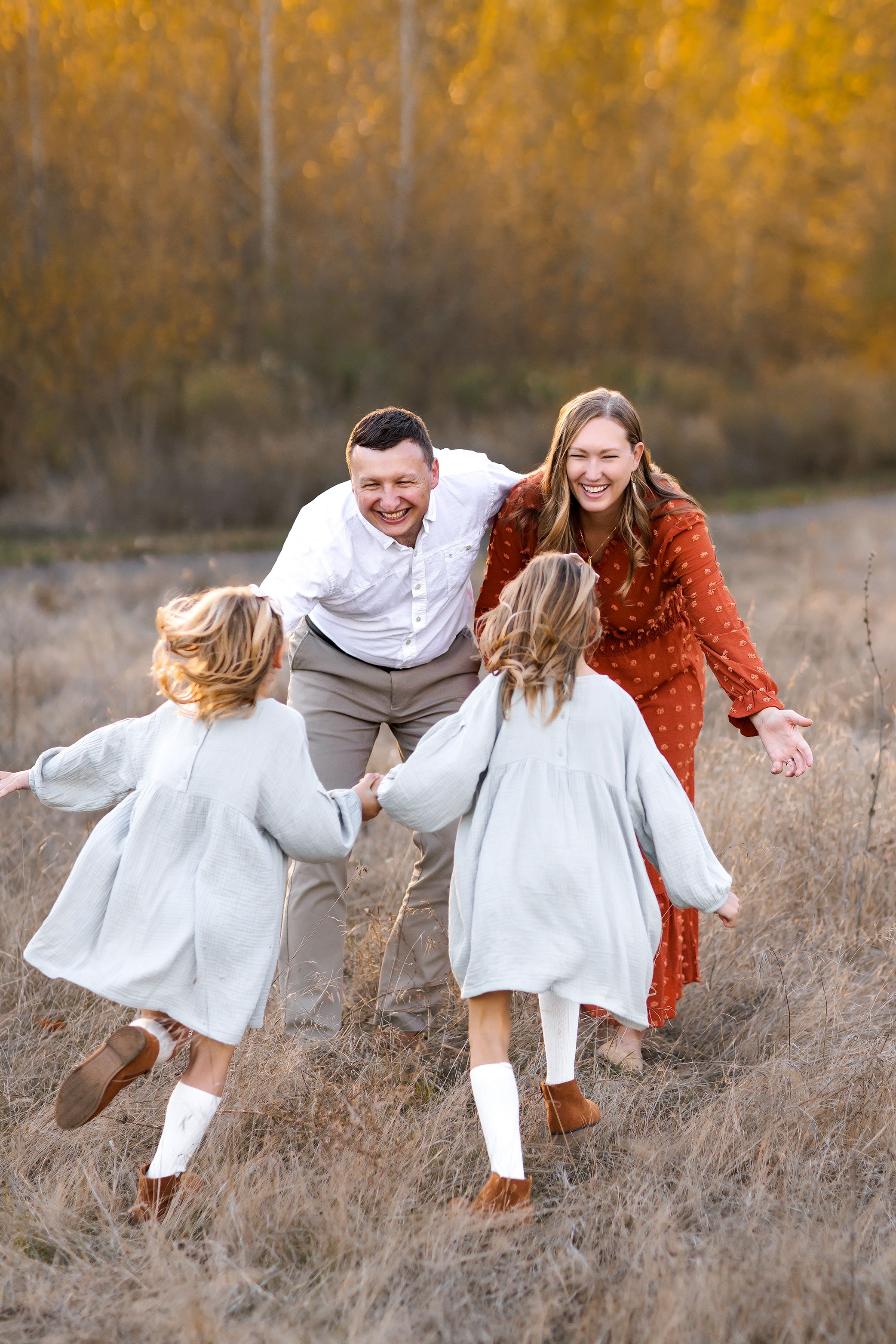 Two young girls run toward their smiling parents in a golden field at sunset during a playful family session.