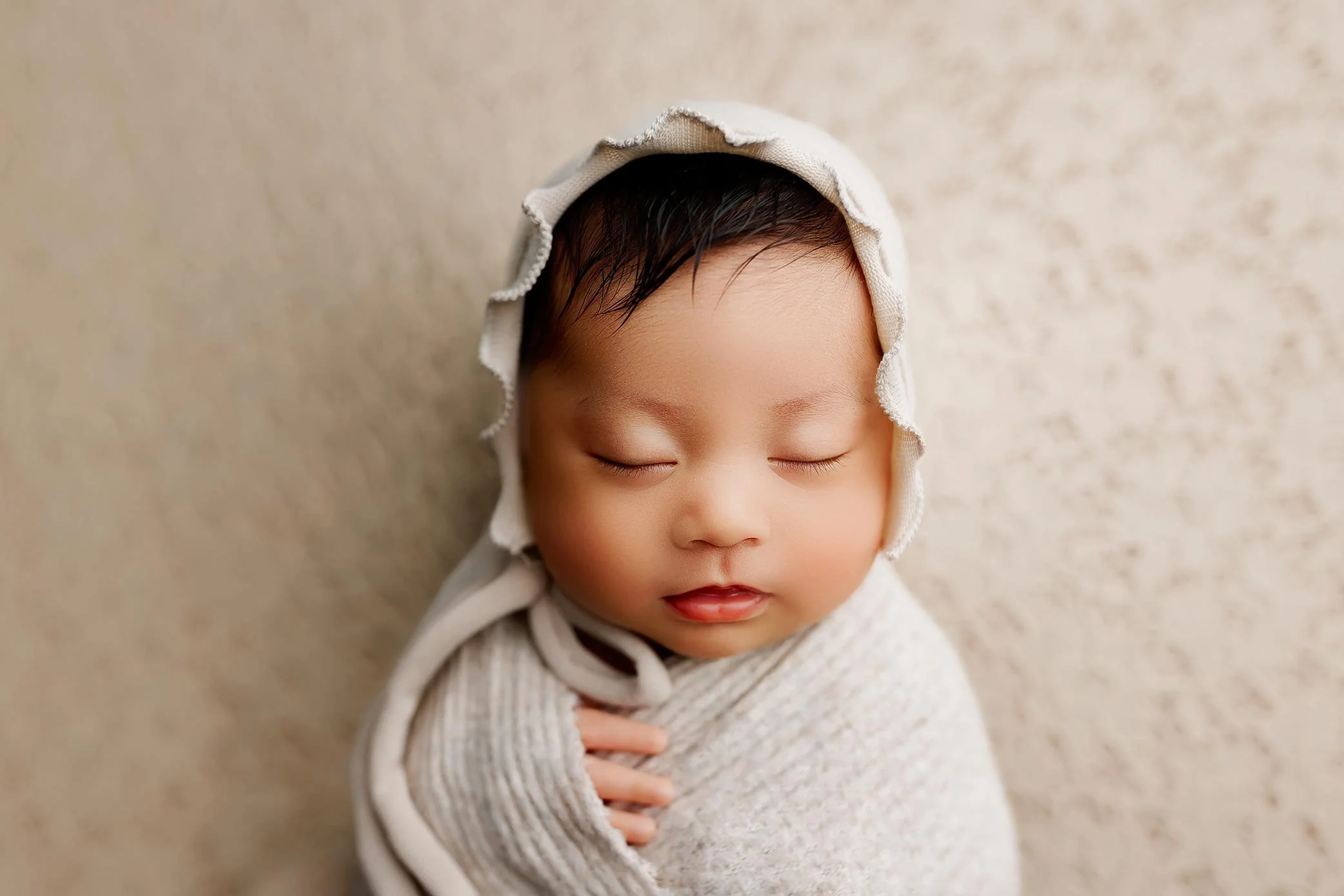 Sleeping newborn wrapped in soft neutral fabric wearing bonnet during posed studio newborn session.