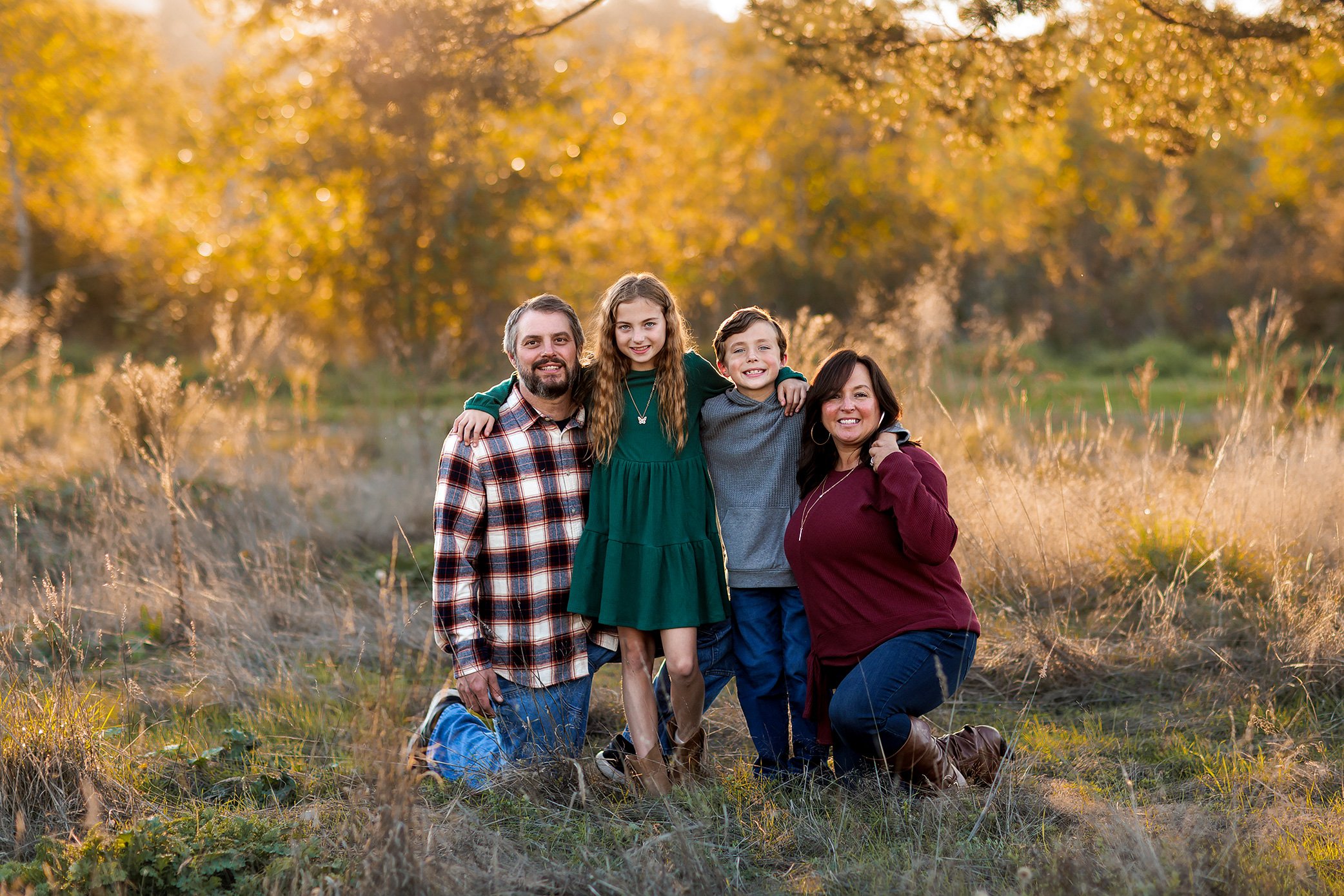 Family of four kneeling together in golden field at sunset with children standing between parents.