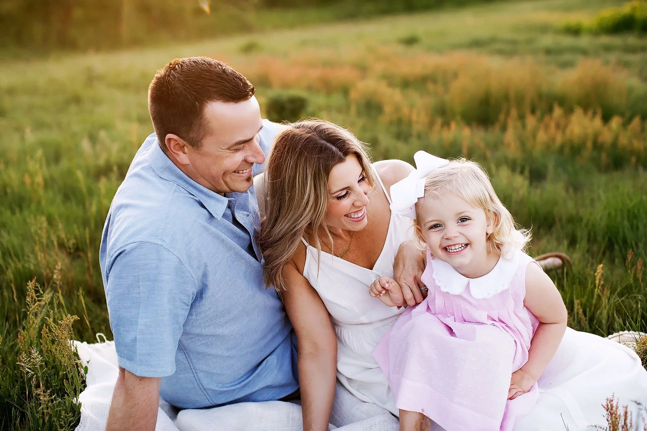 Parents hugging young daughter closely in grassy field at sunset