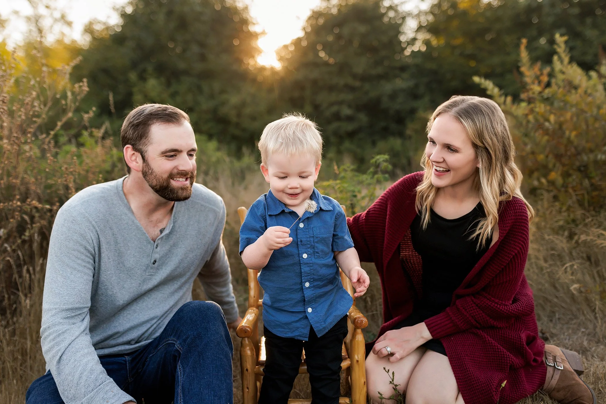 Parents kneeling and smiling at toddler sitting on chair during golden hour outdoor family session.