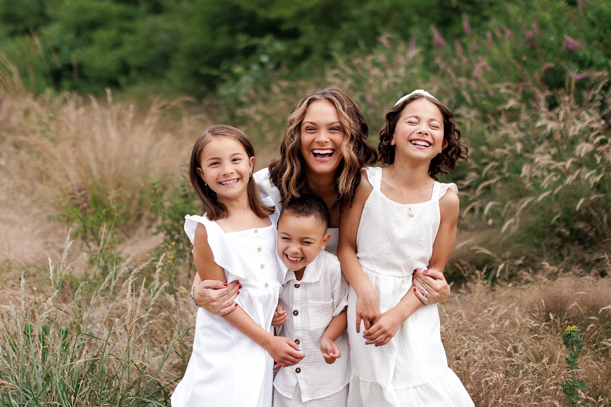 Mother laughing with three children standing together in tall grass.