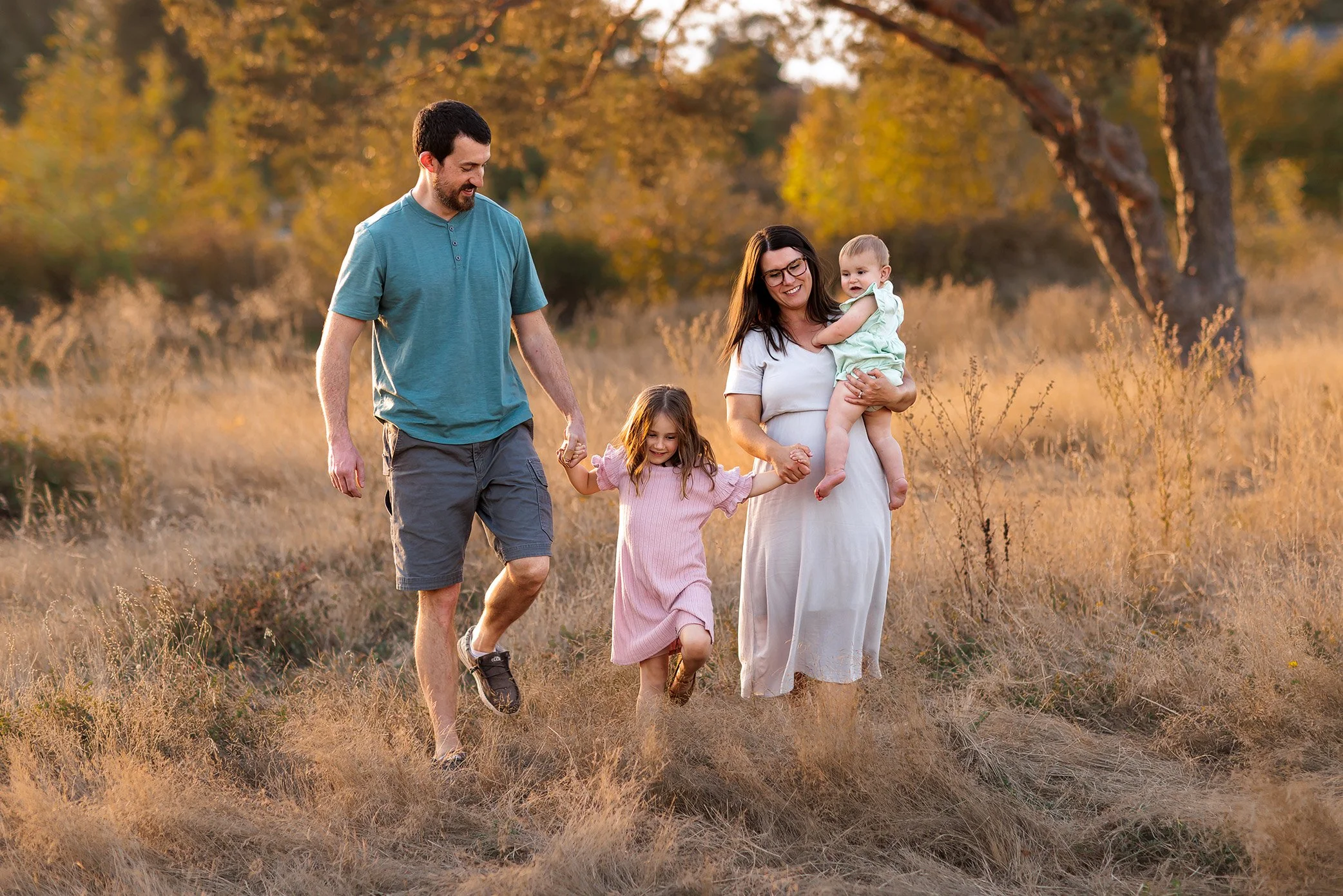 Family of four walking hand in hand through golden field with parents holding toddler and baby.