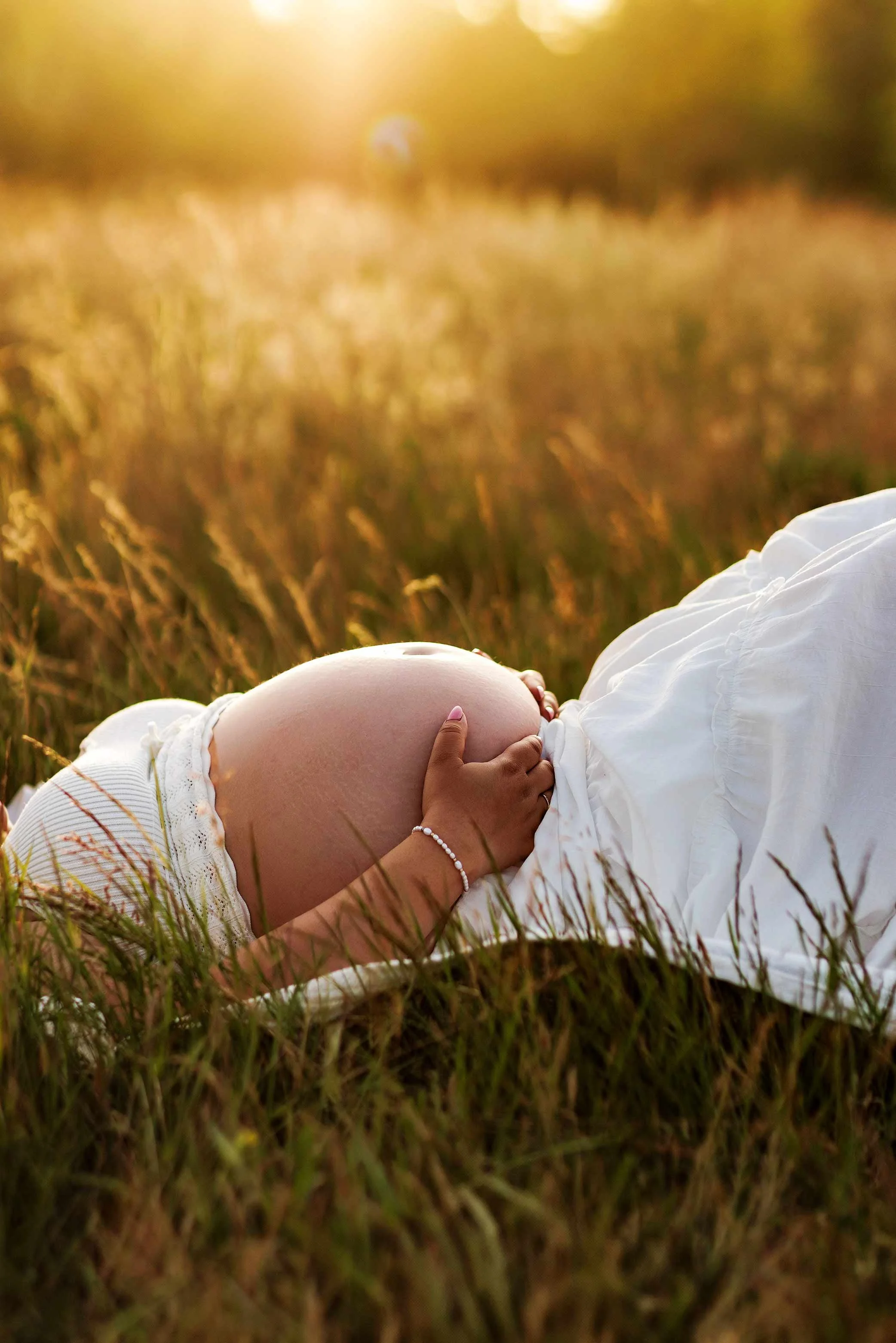 Close-up of pregnant belly in tall grass at sunset, soft golden hour maternity detail