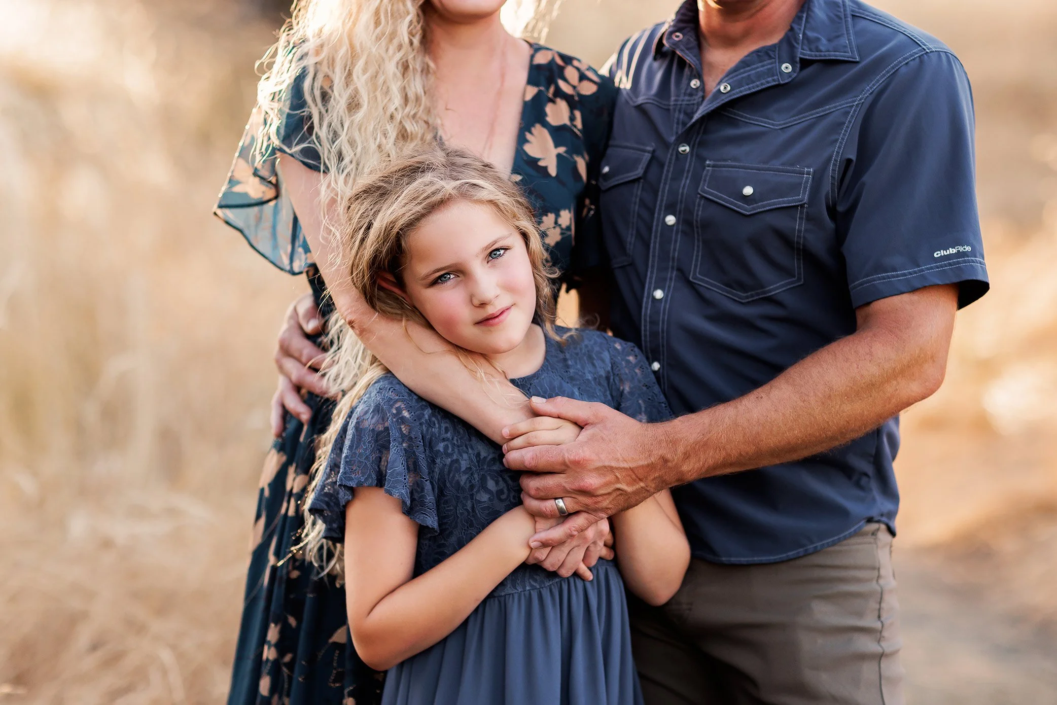 Close up of parents hugging older daughter during outdoor family session in golden light.