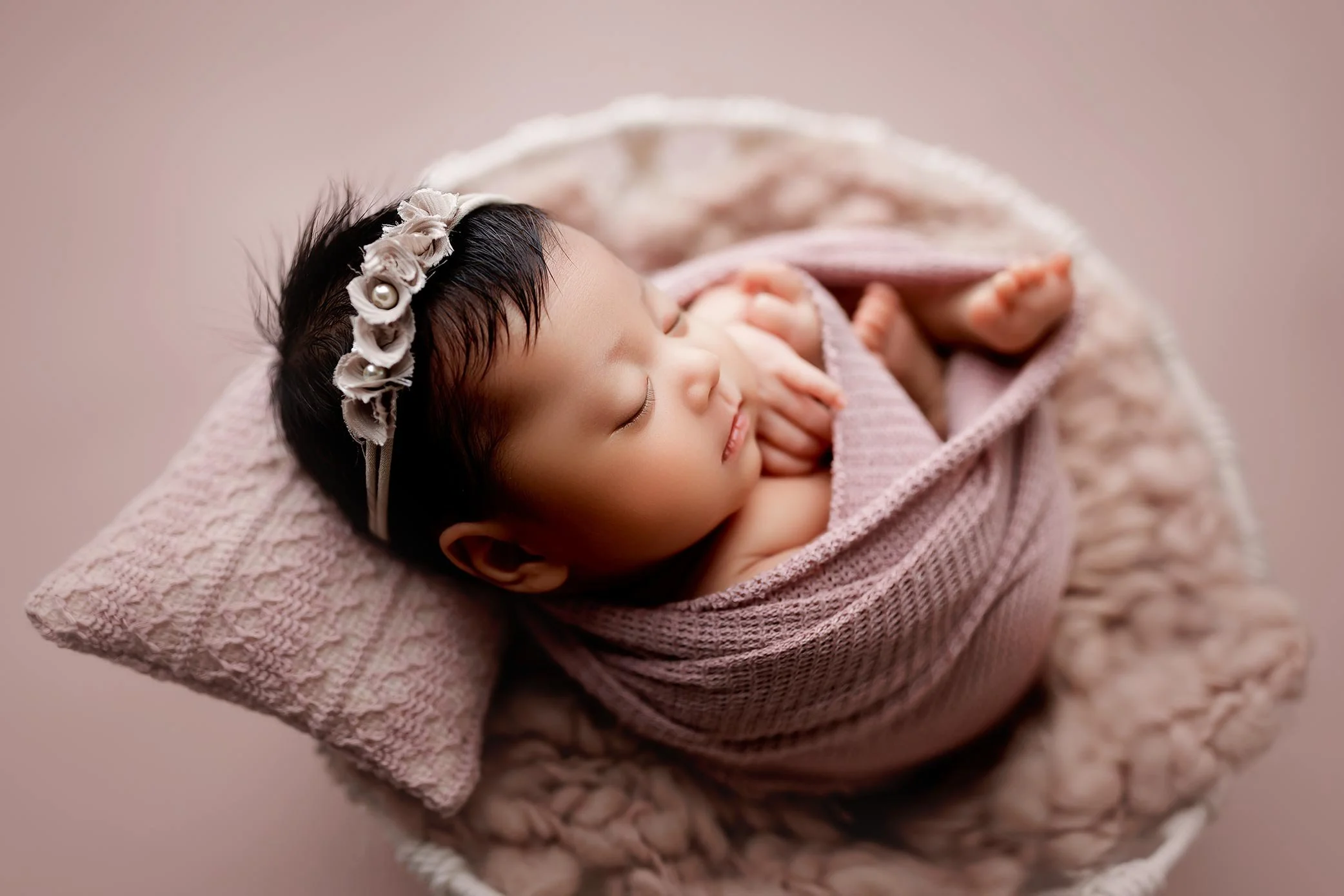 Wrapped newborn posed in a pink wooden bowl with matching wrap, and flower headband.