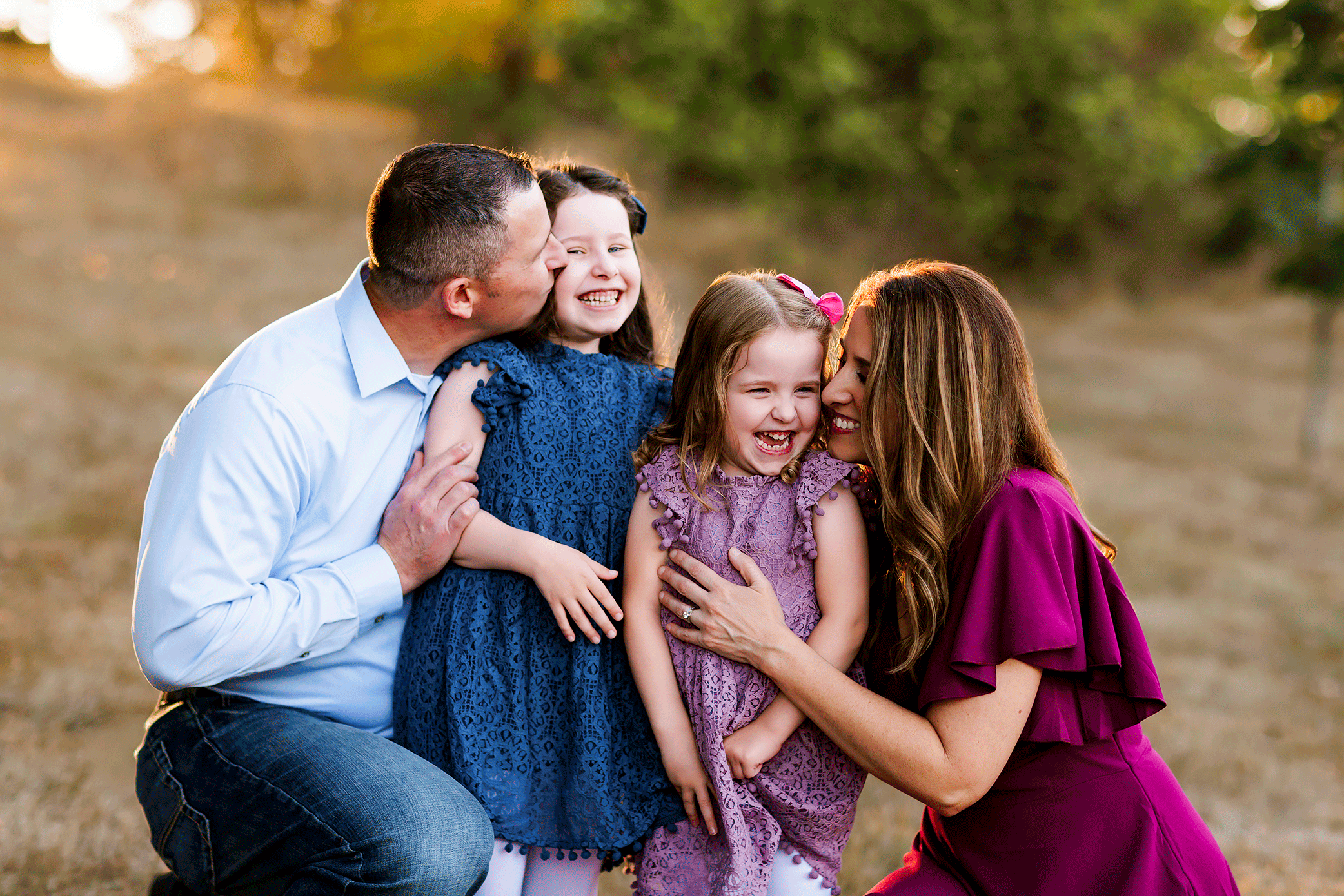 Family of four crouched close together hugging and laughing in golden field.