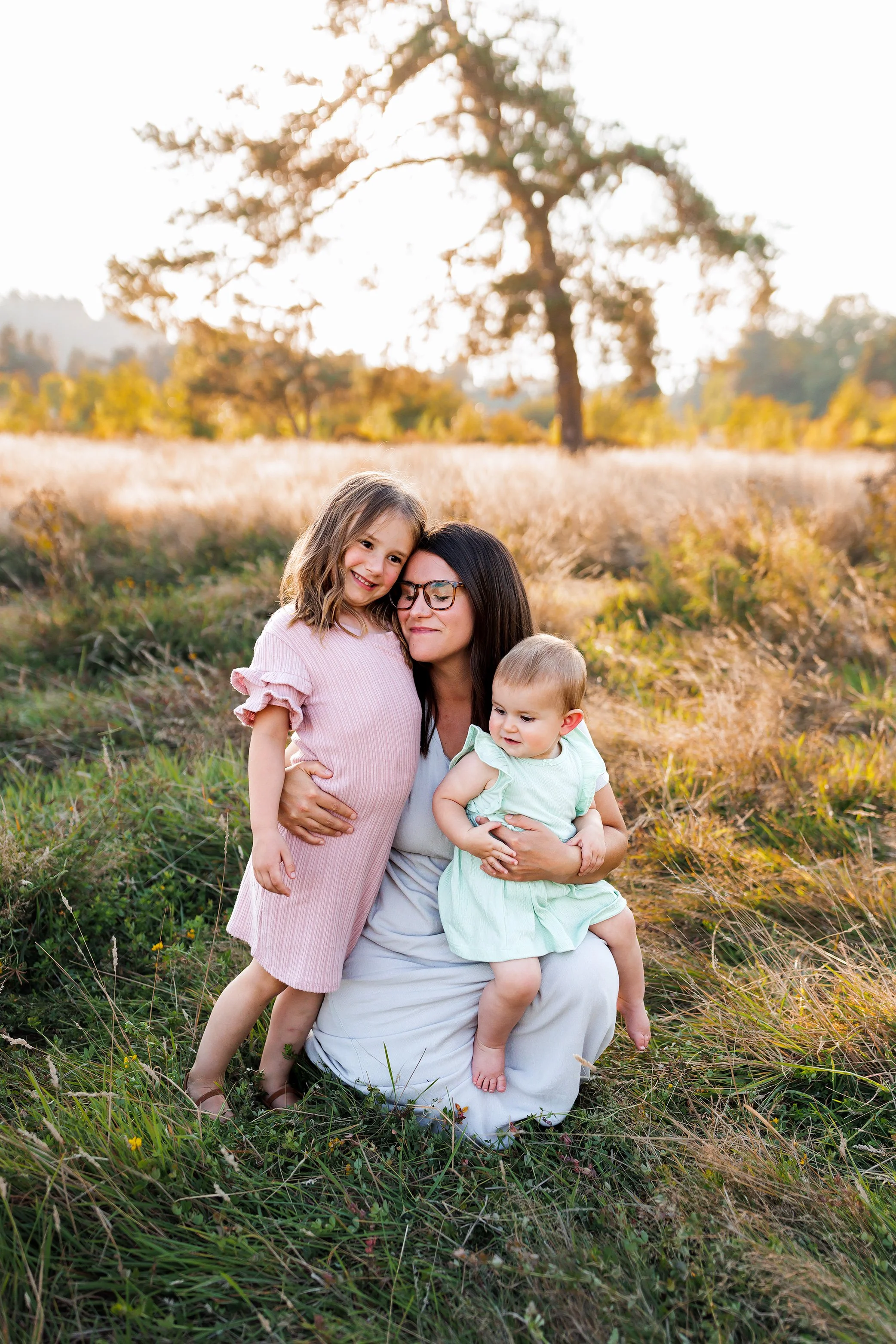 Mom kneeling in field holding baby on her knee while hugging toddler daughter close.