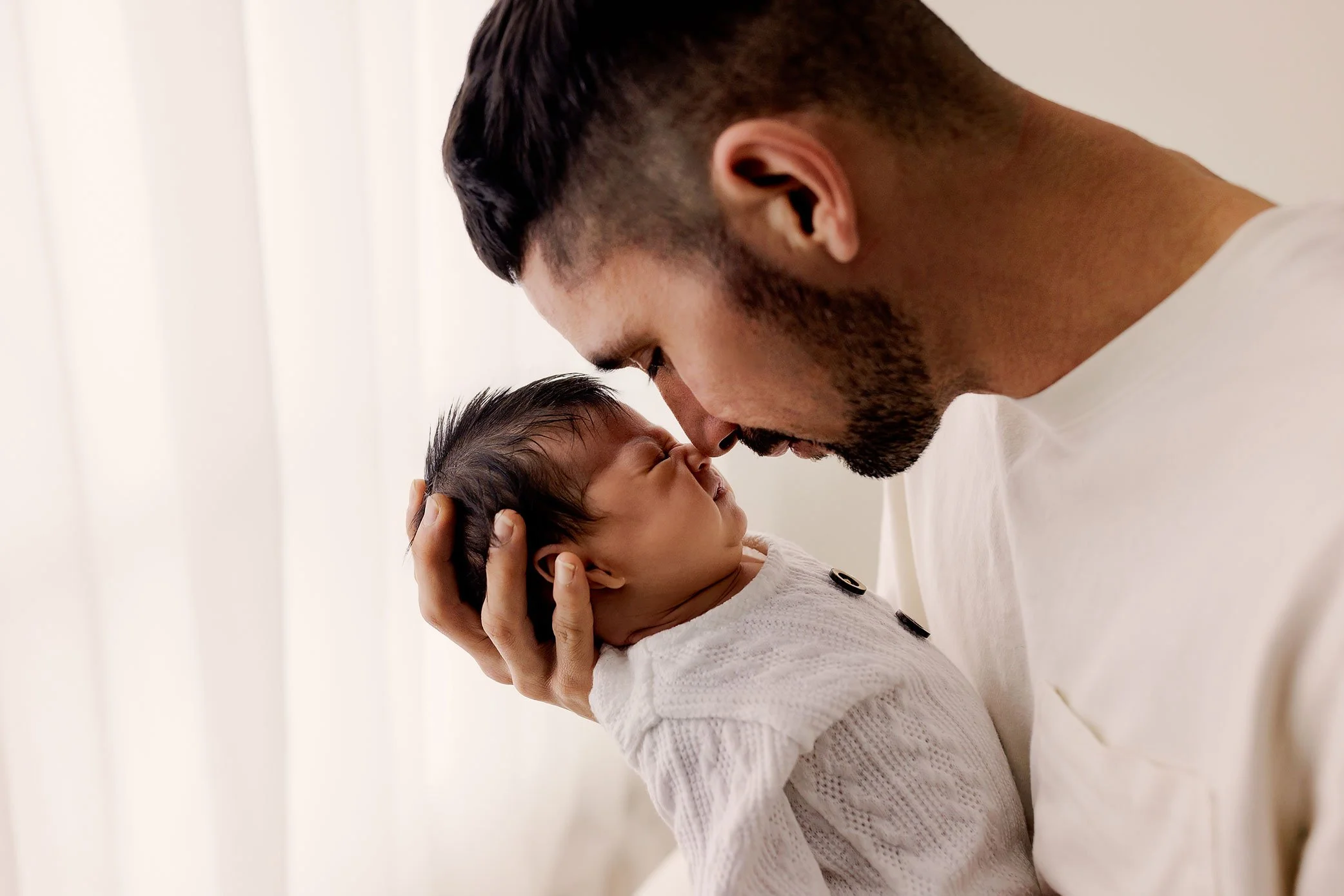 Father gently touching noses with his newborn baby in soft natural window light.