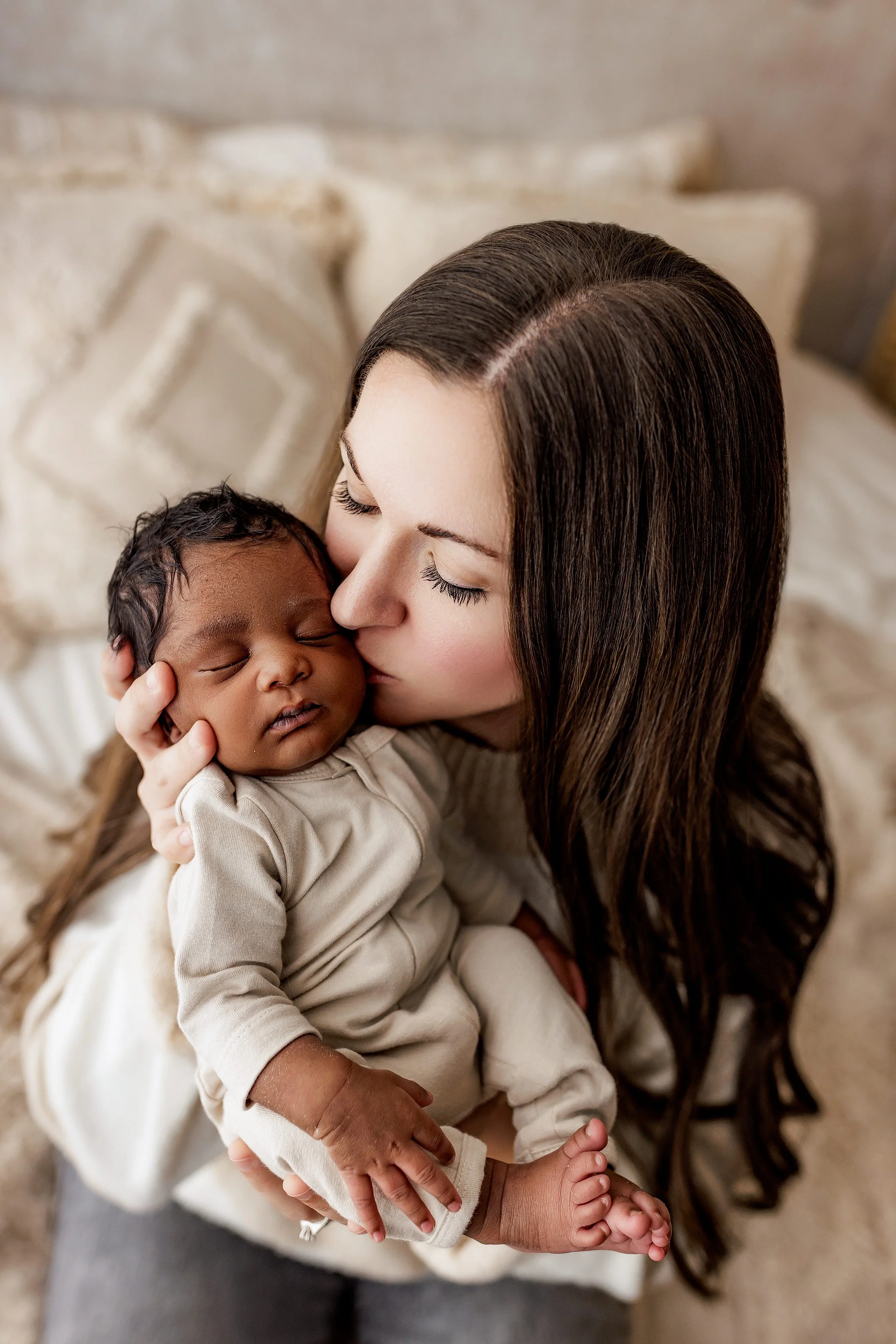 Mother kissing her newborn baby’s cheek while holding him in a soft neutral home setting.