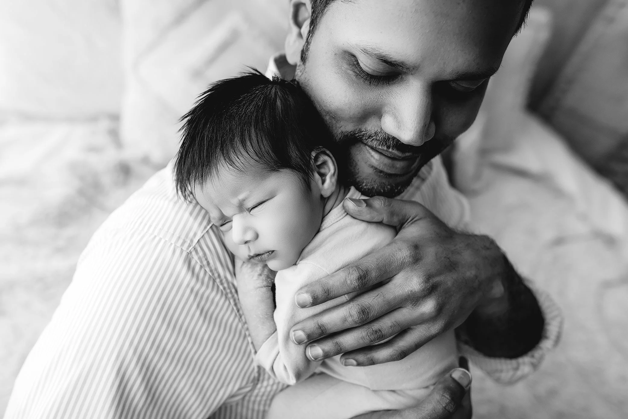 Black and white image of baby sleeping on his father’s chest, in a little outfit.