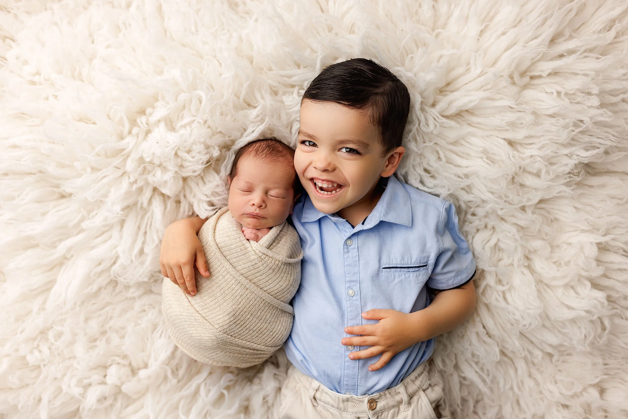 Toddler sibling smiling while cuddling a swaddled newborn during a Salem, Oregon newborn photography session on a soft neutral backdrop.