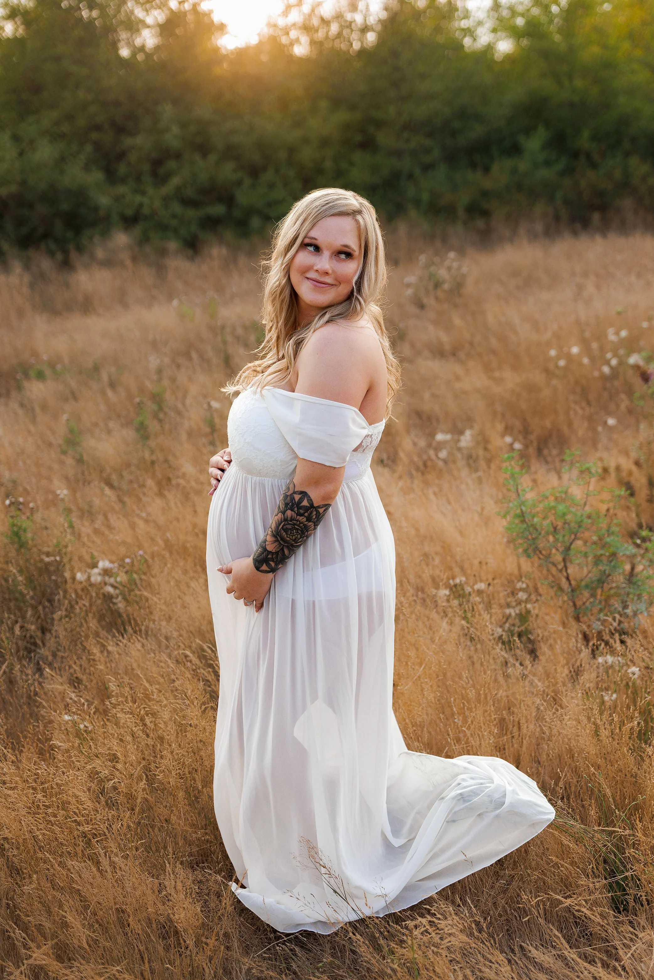Expecting mother looking over her shoulder in a flowing white dress standing in golden field at sunset maternity session Salem Oregon