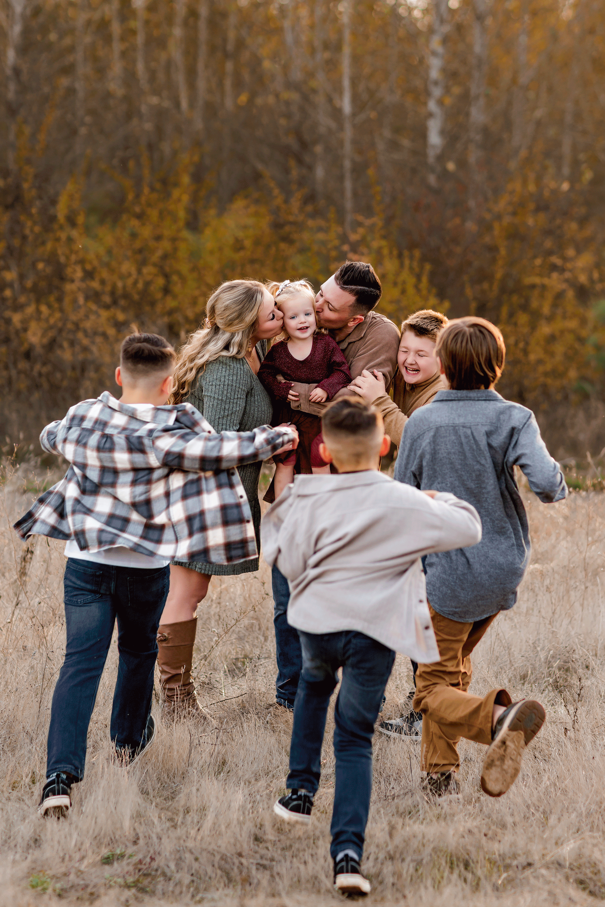 Large family gathers around parents holding a toddler, sharing hugs and laughter in a fall field at sunset.