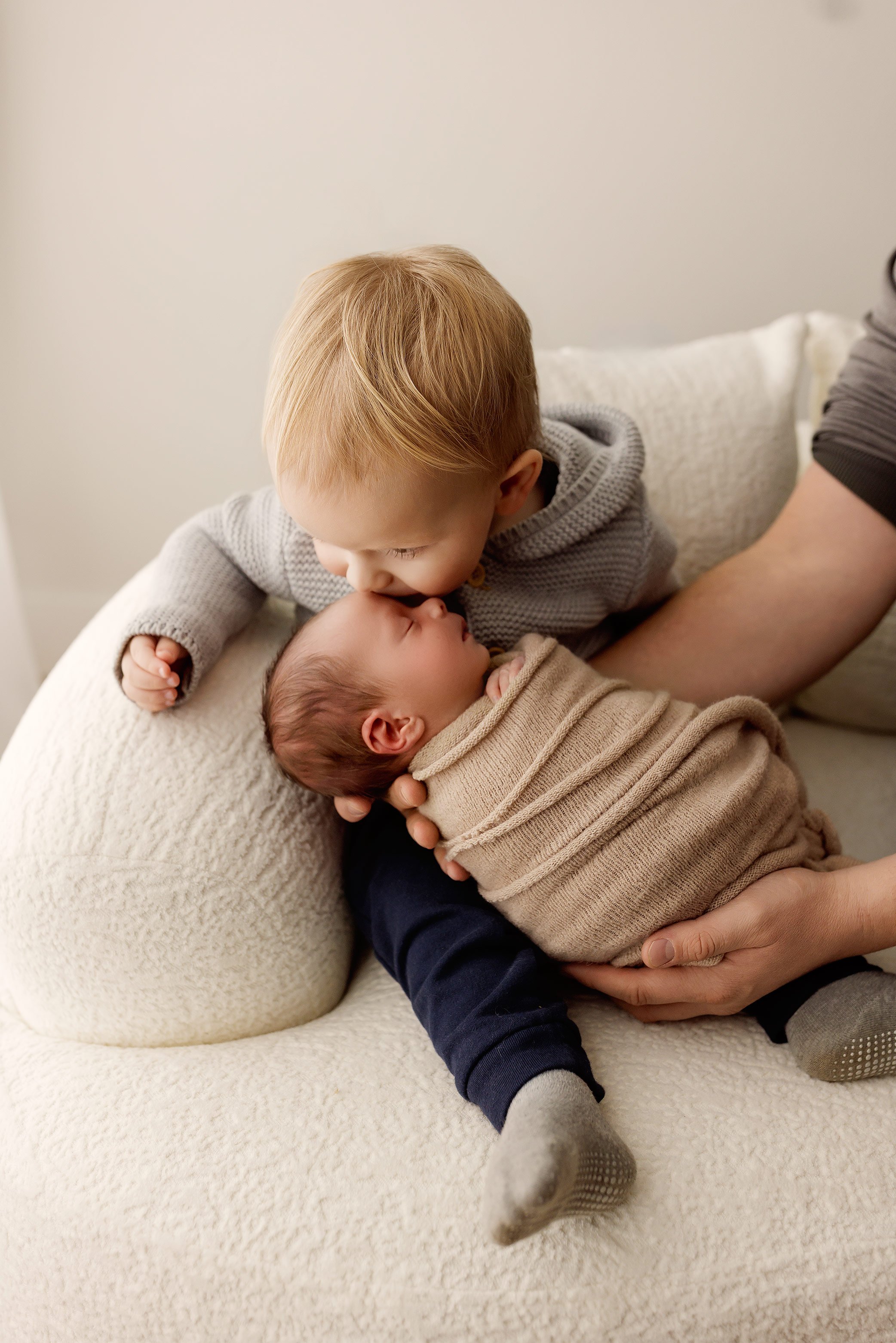 Older sibling gently kissing a newborn baby wrapped in a soft neutral blanket