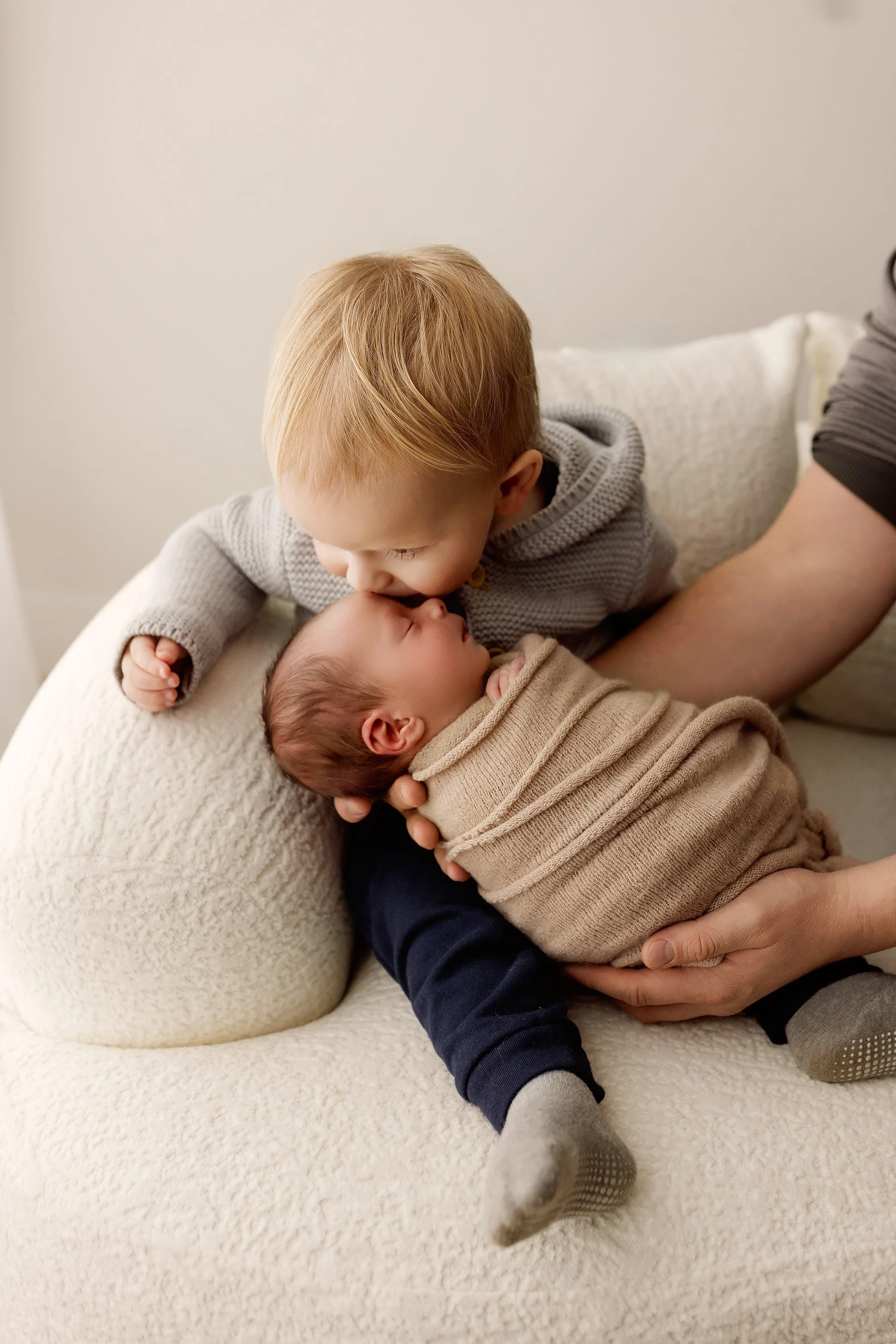 Toddler gently kissing newborn baby while dad's hands support baby.