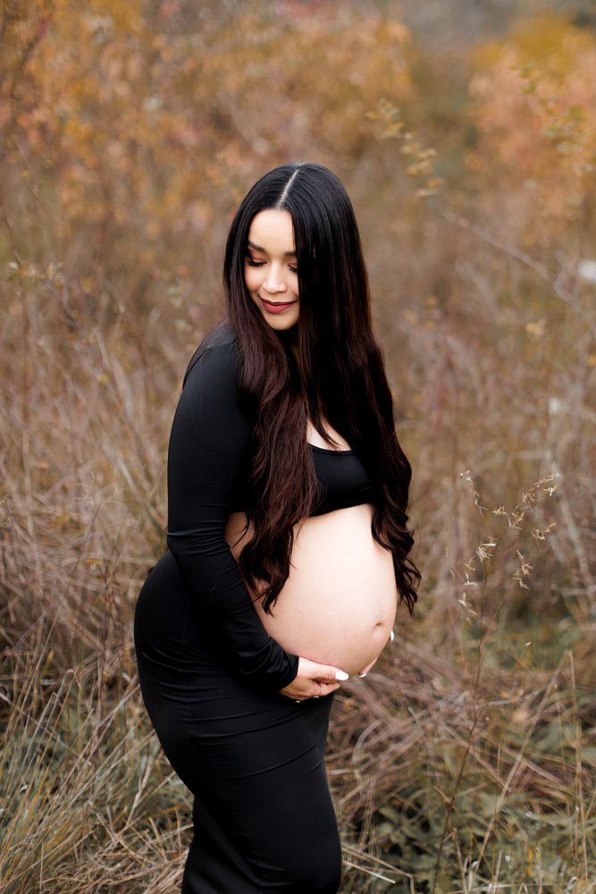 Pregnant mother in black dress holding belly in natural outdoor setting during maternity session