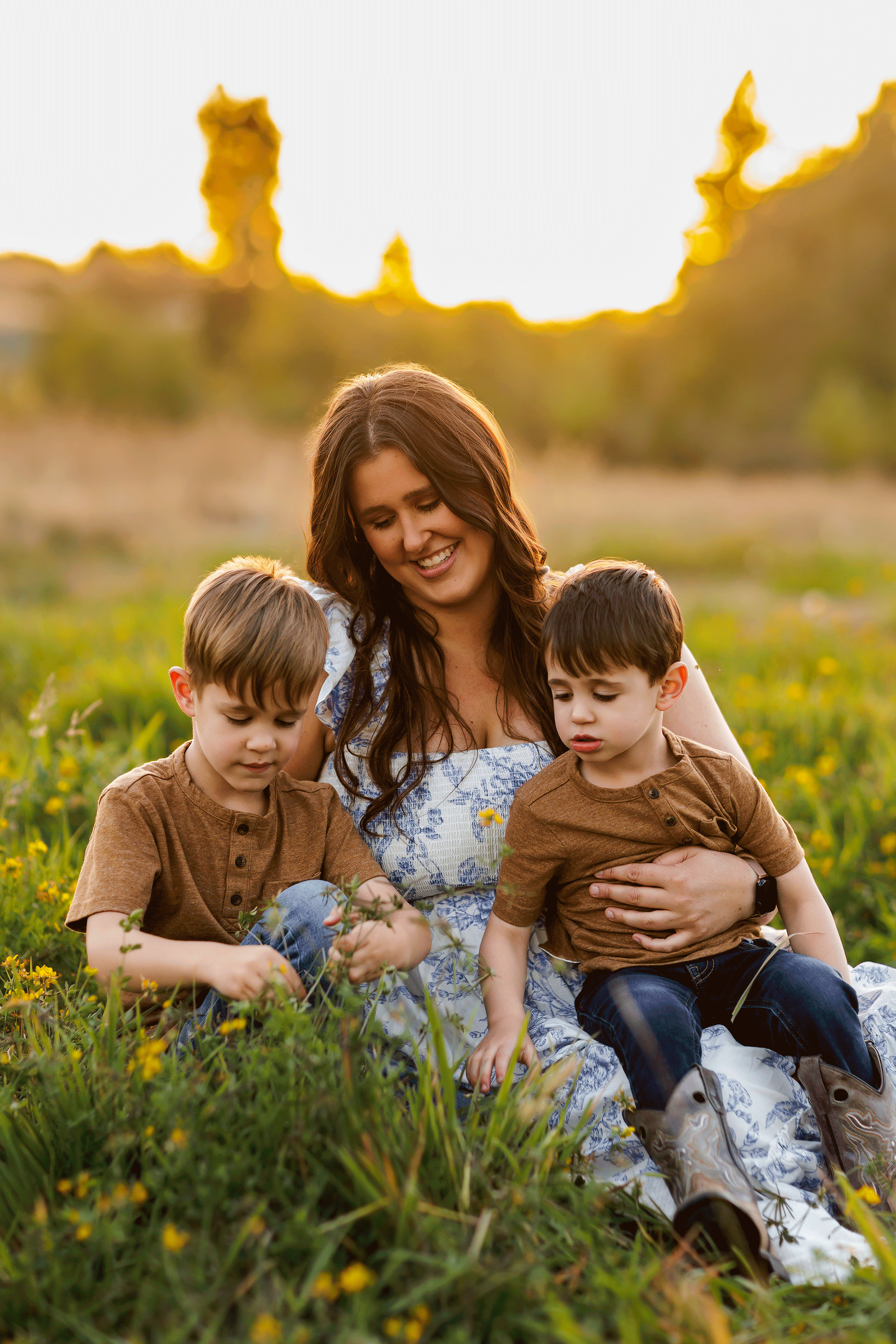 Mother sitting in wildflowers with her two boys exploring grass at sunset