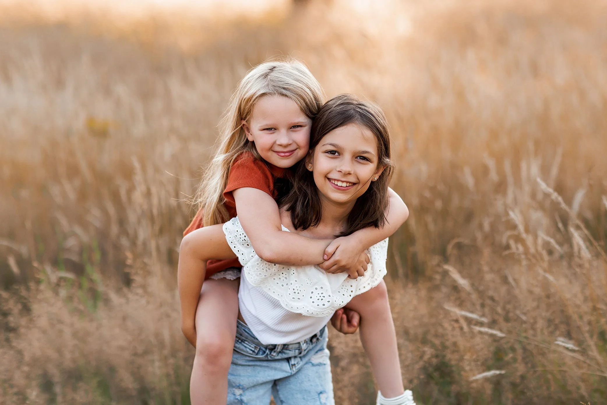 Older sister giving younger sister a piggyback ride in a golden field at sunset