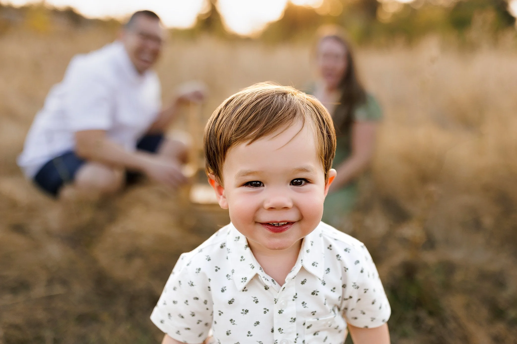 Toddler smiling in foreground with parents softly blurred in background during golden hour family session.