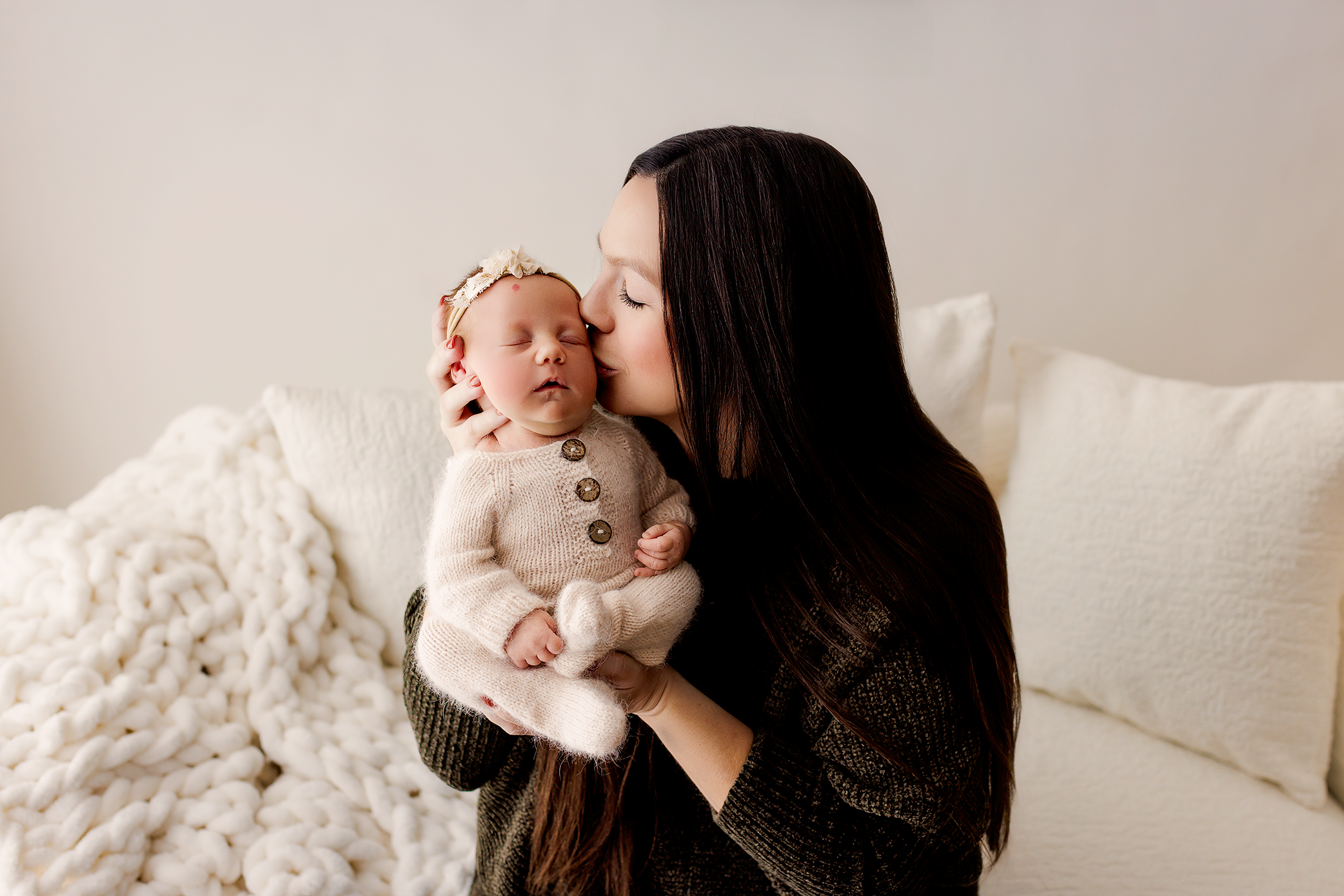 Mother kissing sleeping newborn baby dressed in cream knit outfit while seated on bed.