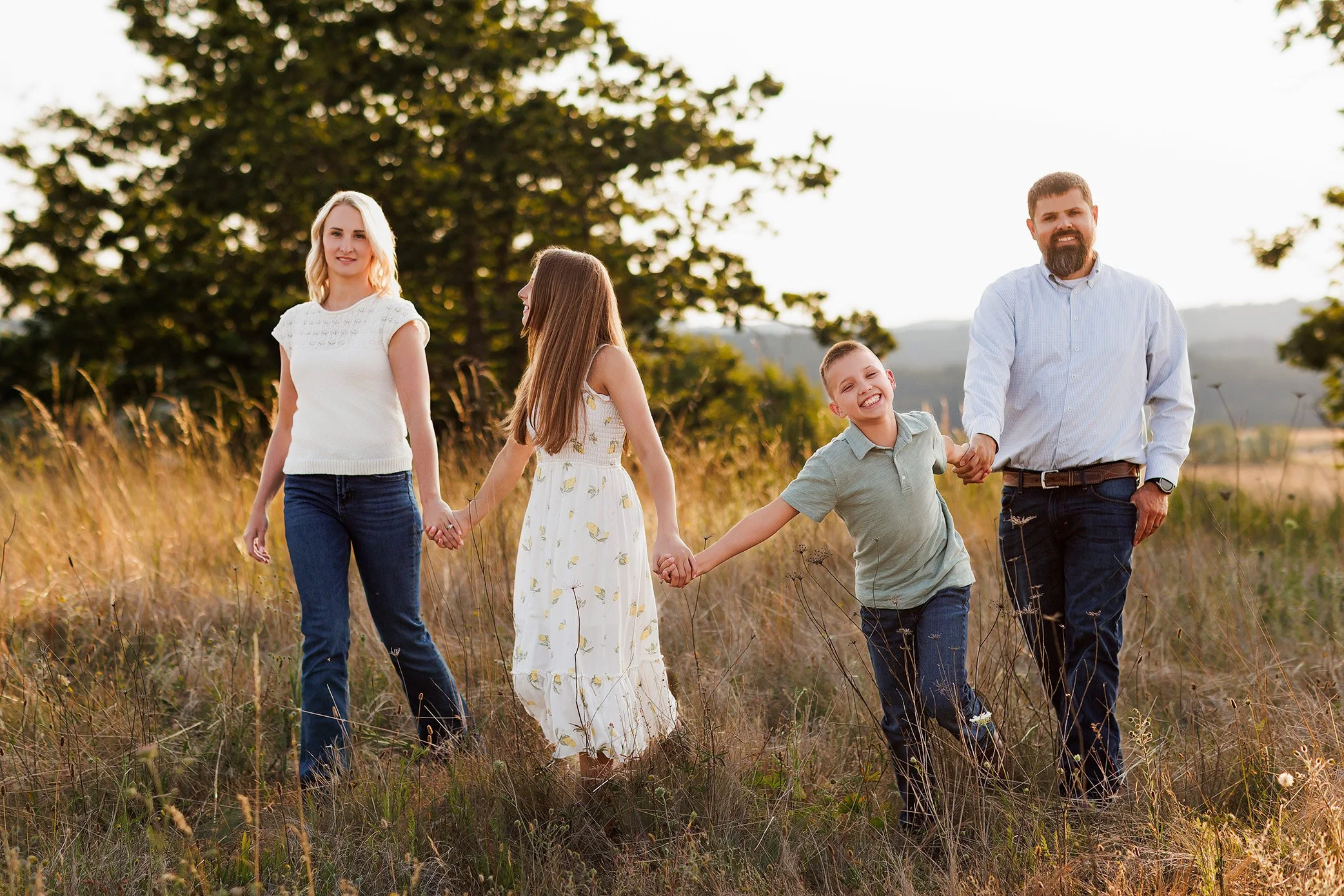 Family of four holding hands walking through tall grass at golden hour