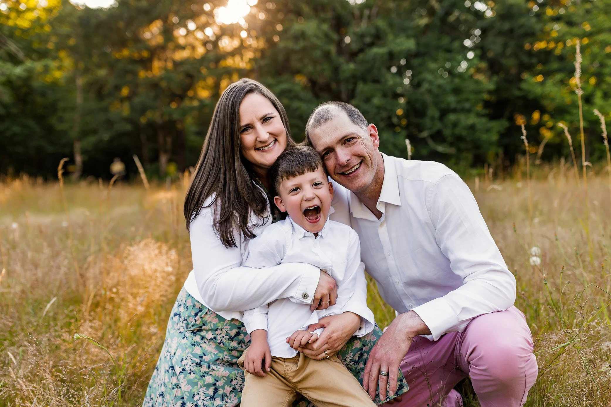 Family of three sitting close together in golden field during outdoor photography session in Salem Oregon