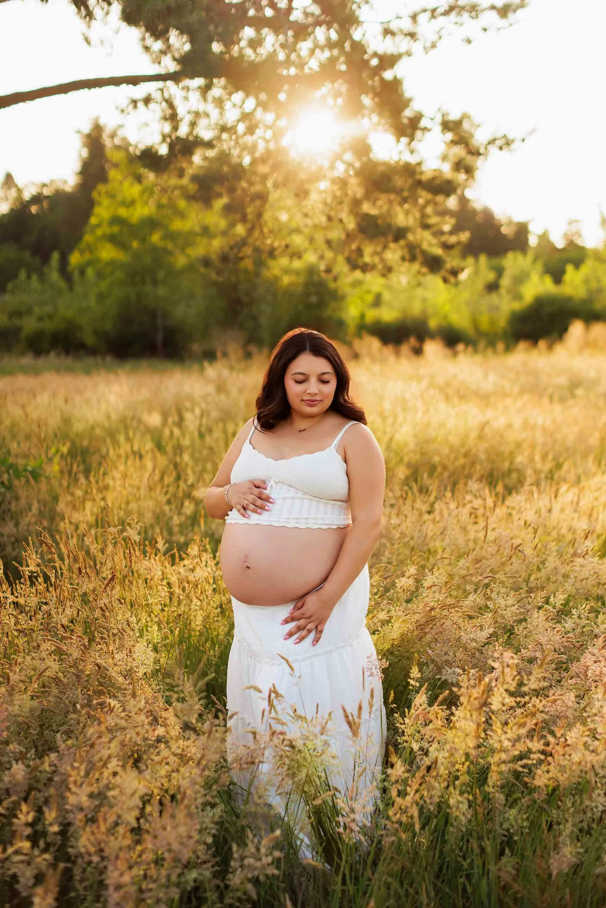Pregnant woman in white dress standing in field at sunset, natural light maternity portrait