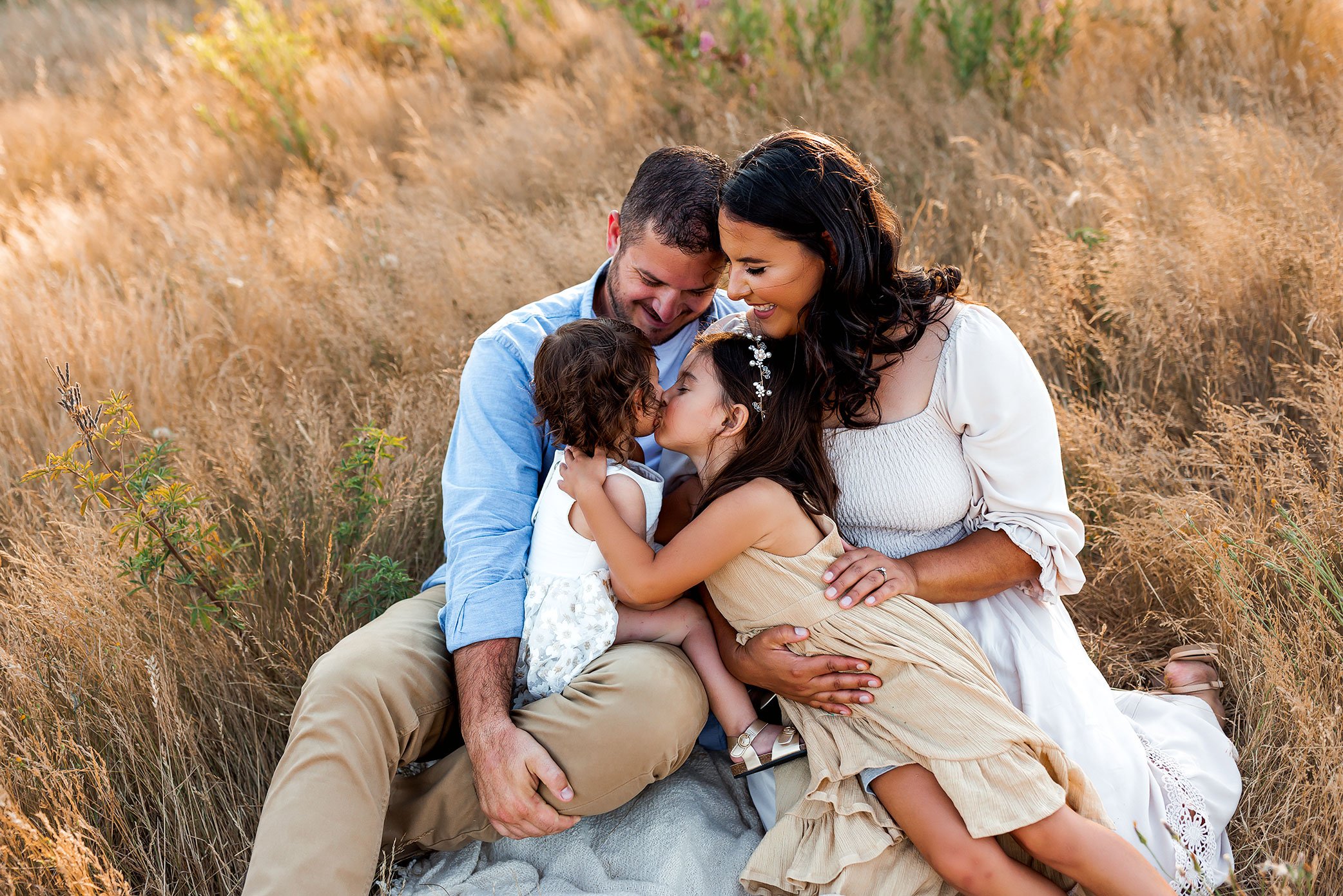 Family sitting closely together in tall golden grass with sisters hugging