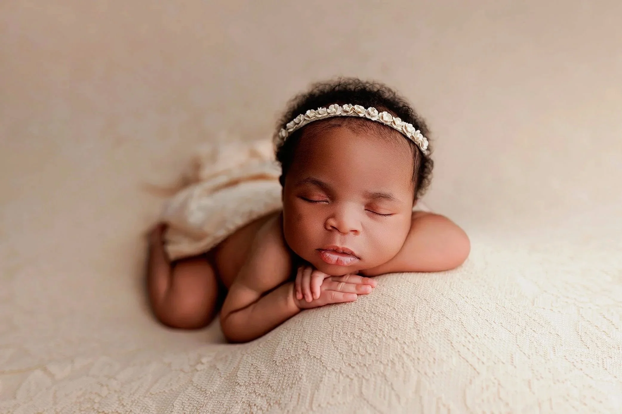 Infant baby laying posed on a cream blanket with a neutral floral headband.