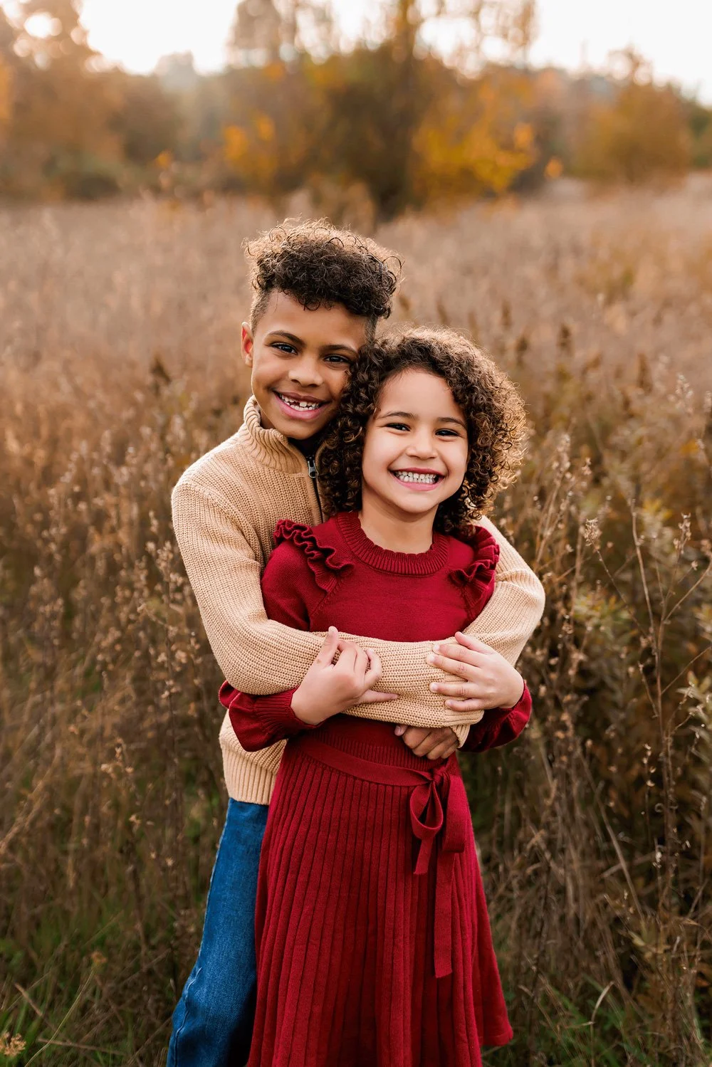 Brother and sister smiling and hugging in a golden autumn field, wearing cozy sweaters during an outdoor photo session.