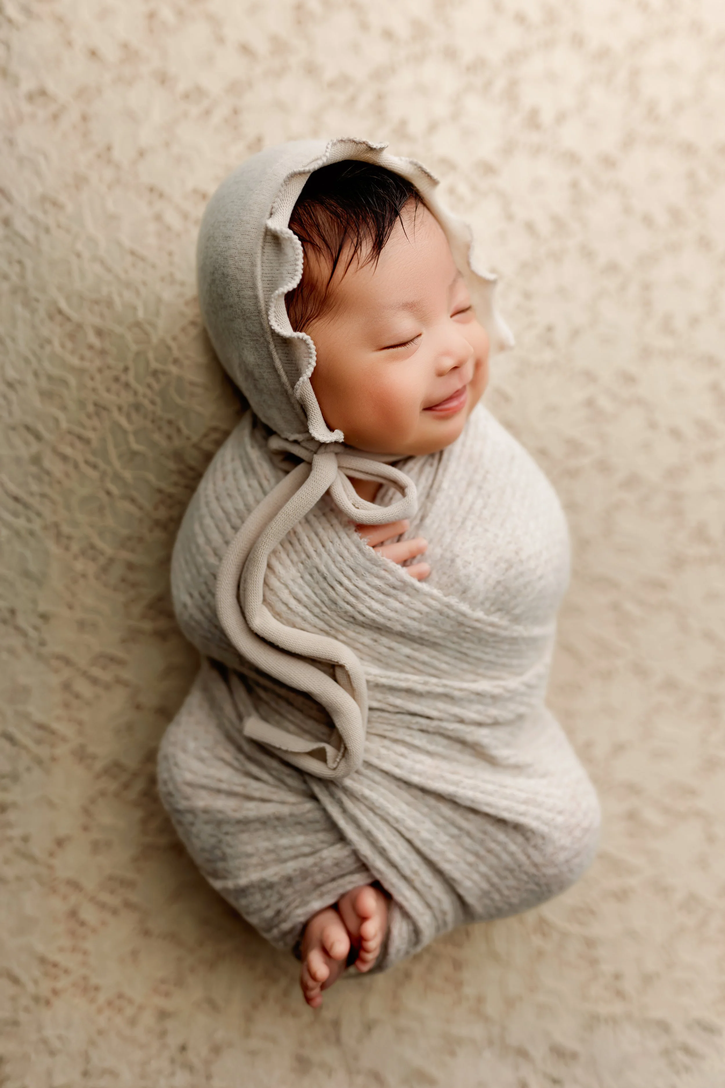 Sleeping newborn smiling while wrapped and wearing a ruffle bonnet during a posed newborn photography session.