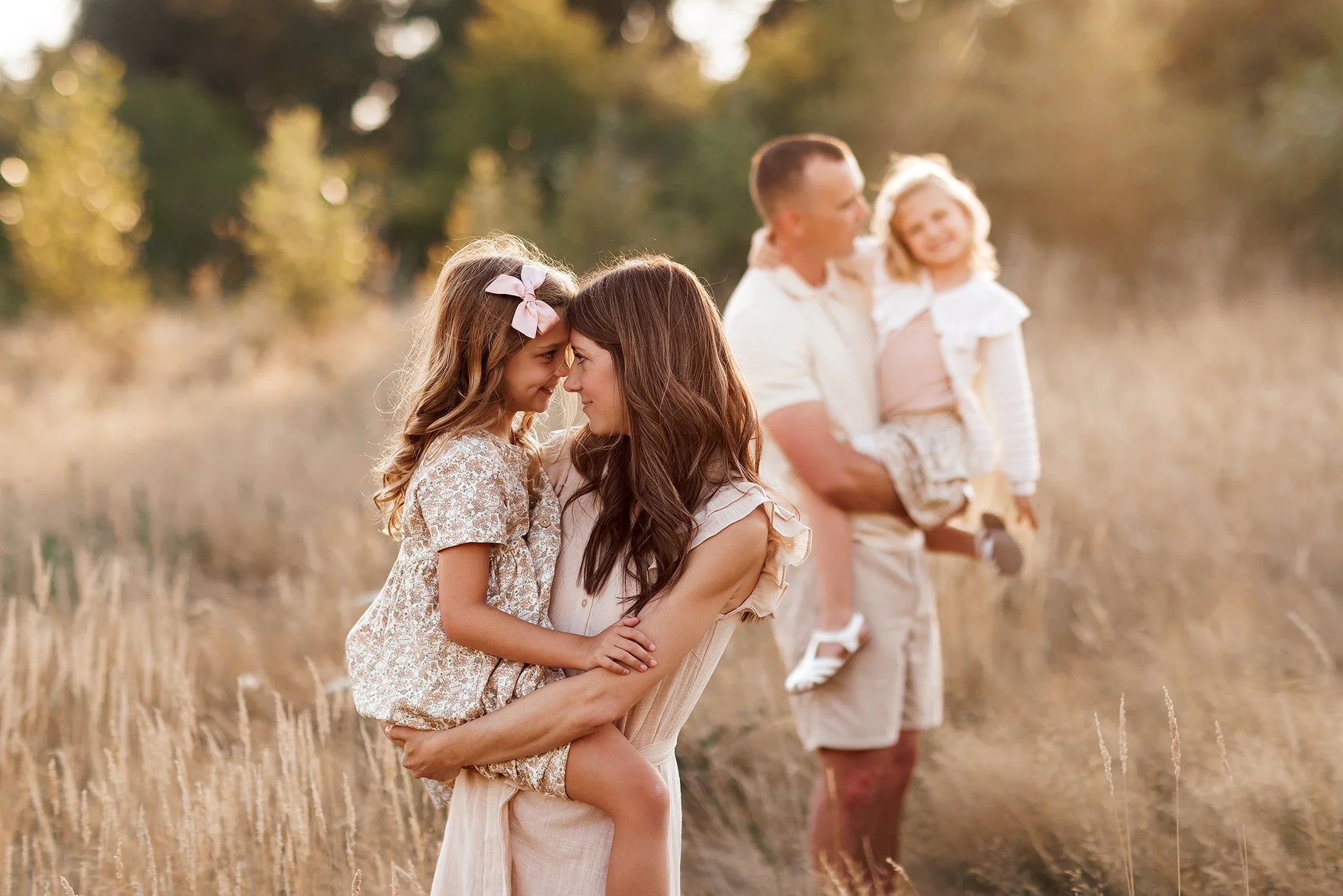Mom holding daughter in sharp focus with dad and sister softly blurred in background during golden hour family session.