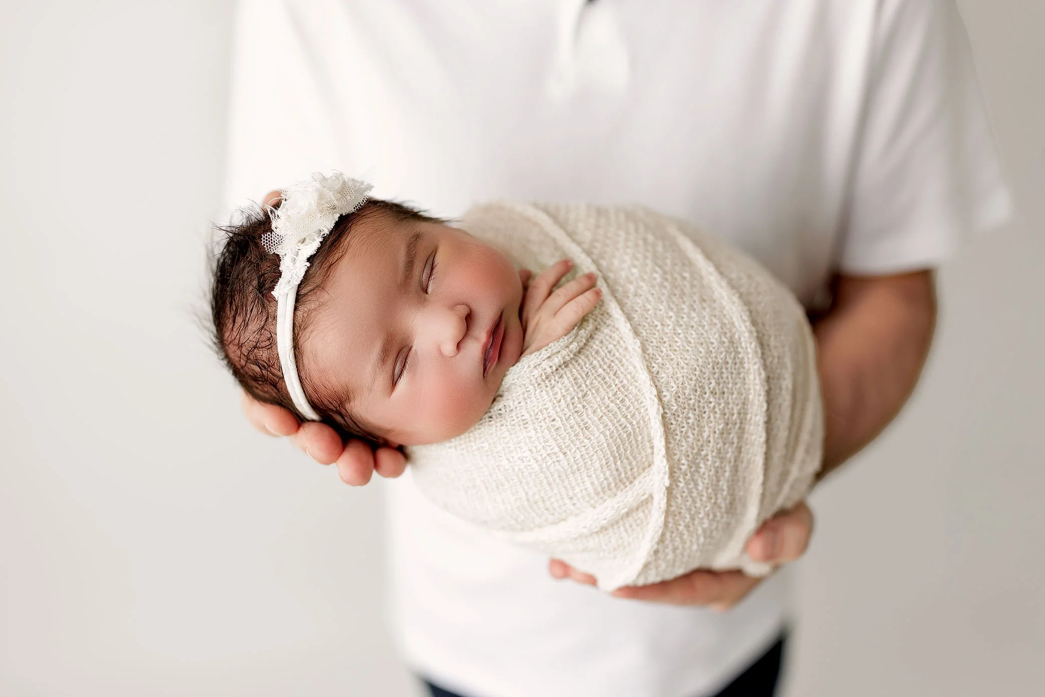 Newborn baby girl wrapped in a cream swaddle, sleeping peacefully in her father’s hands.