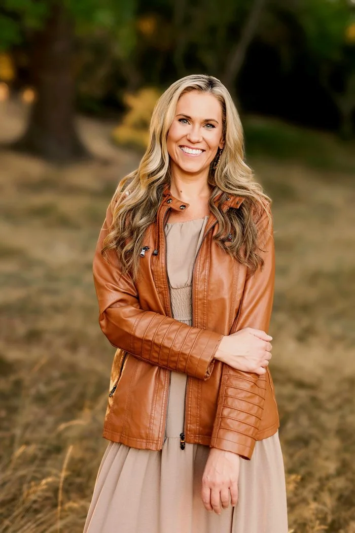 Smiling woman with long blonde hair in tan dress and brown leather jacket, standing outdoors in natural golden light.