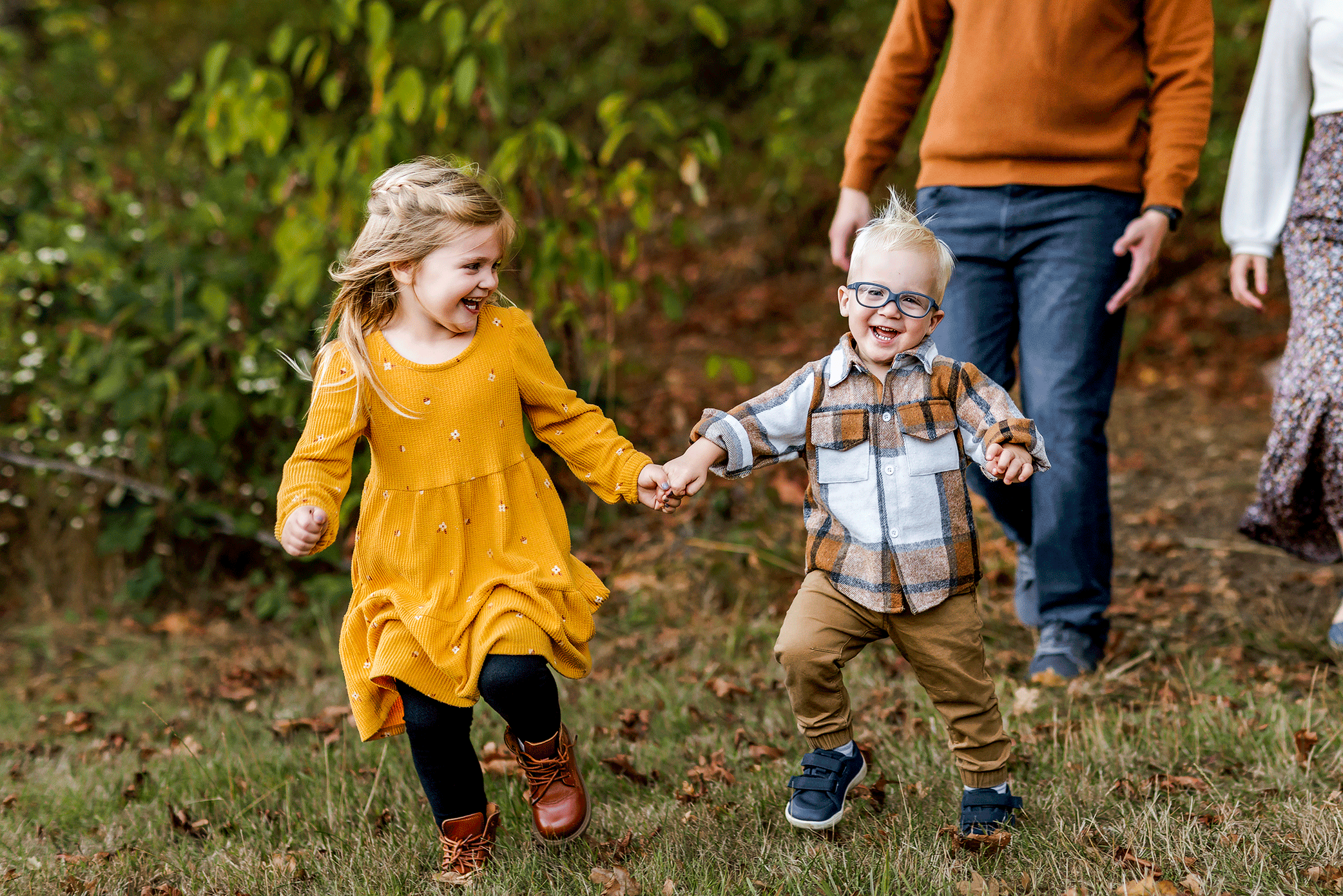Siblings holding hands and running while parents playfully chase behind during outdoor family session.