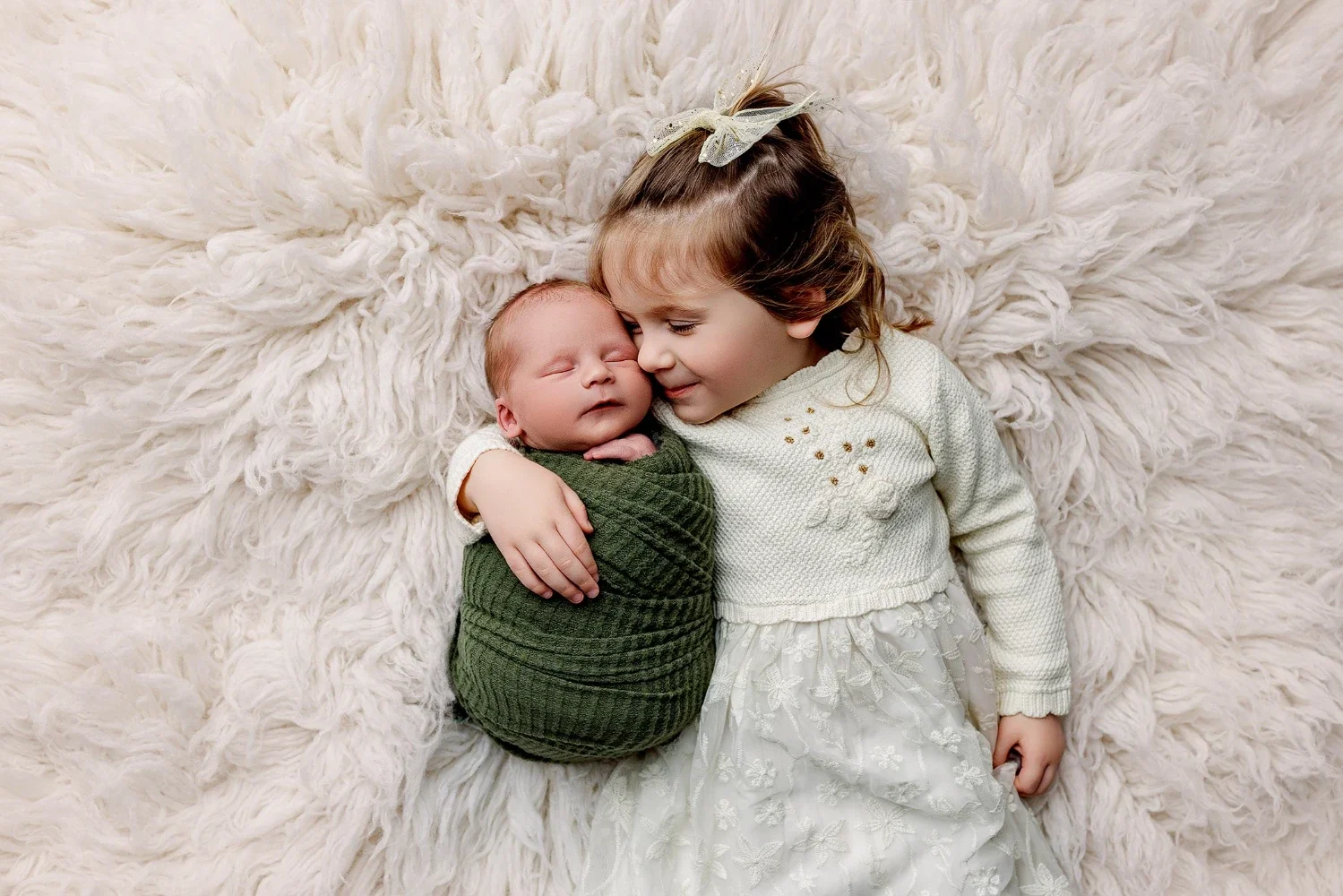 Young sibling gently cuddling newborn baby wrapped in green swaddle on soft neutral blanket, capturing sweet family connection