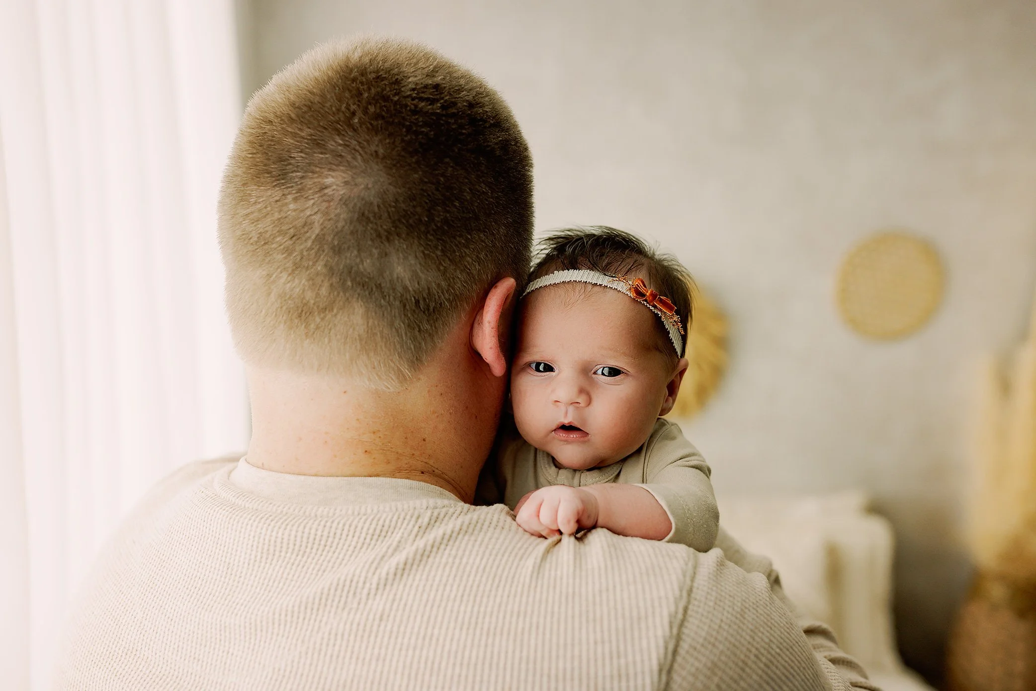 Newborn baby girl resting on her father’s shoulder in a bright, neutral home studio.