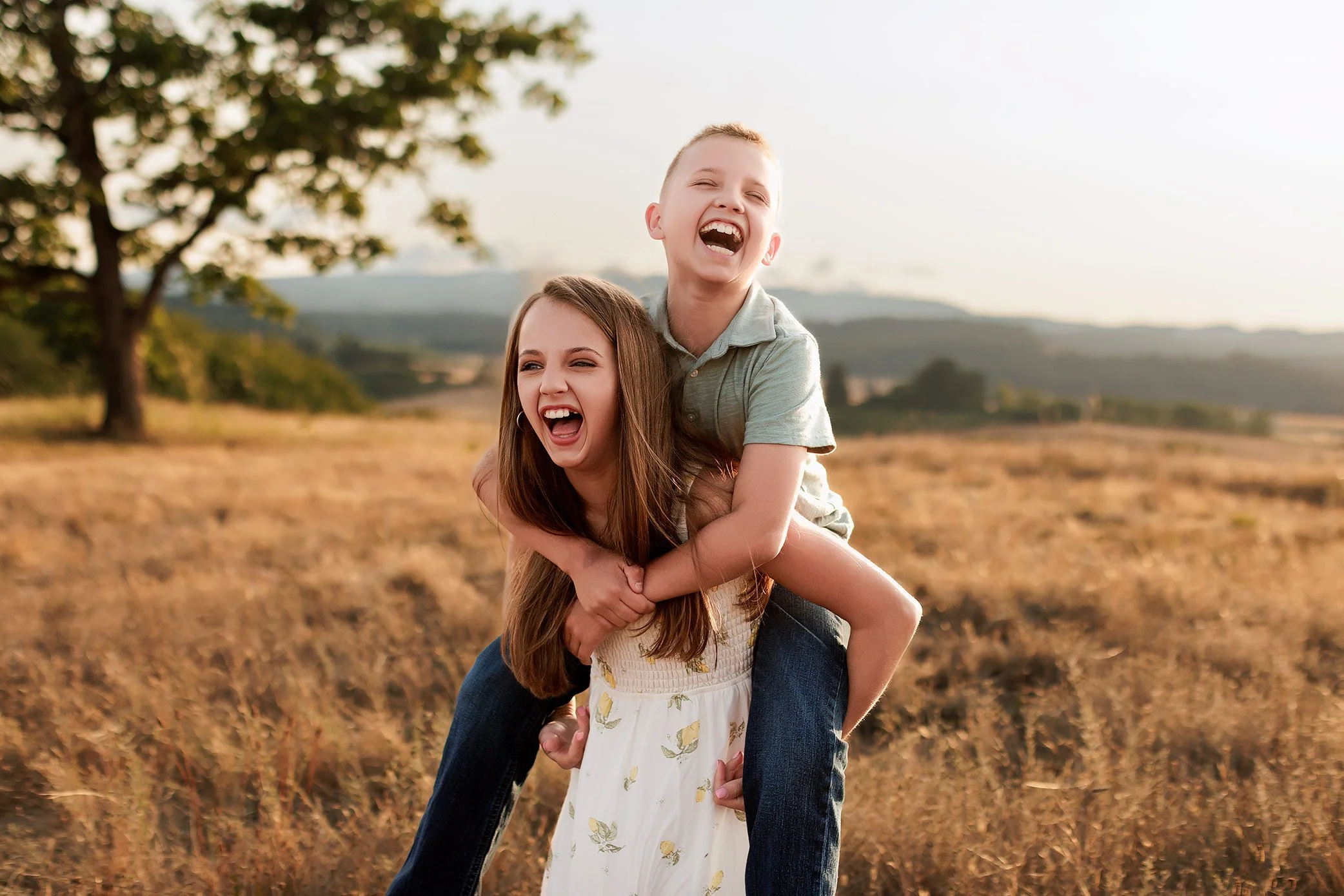 Brother and sister laughing during piggyback ride in golden sunset field