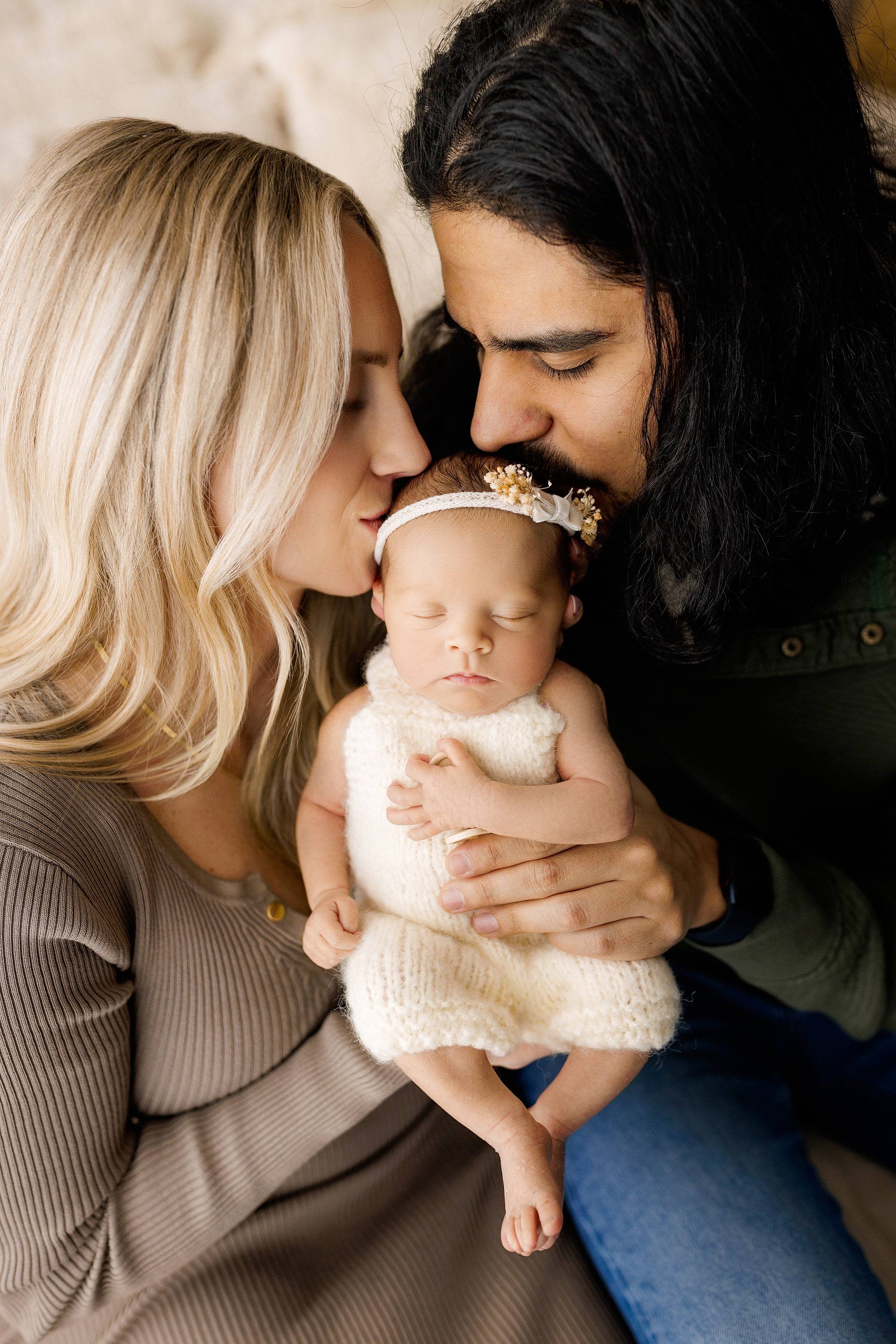 Parents gently kissing their sleeping newborn daughter as they hold her close in warm, soft natural light.